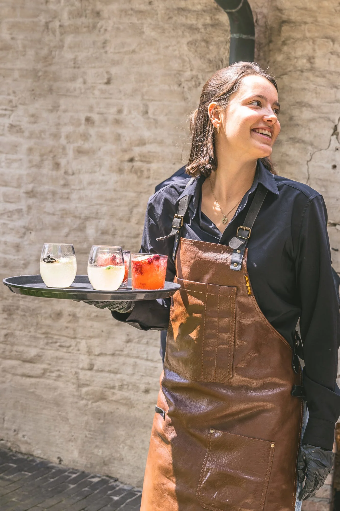 Vrouw met een bruin leren schort die drankjes serveert, waaronder twee witte en een rood drankje, lachend naast een muur.