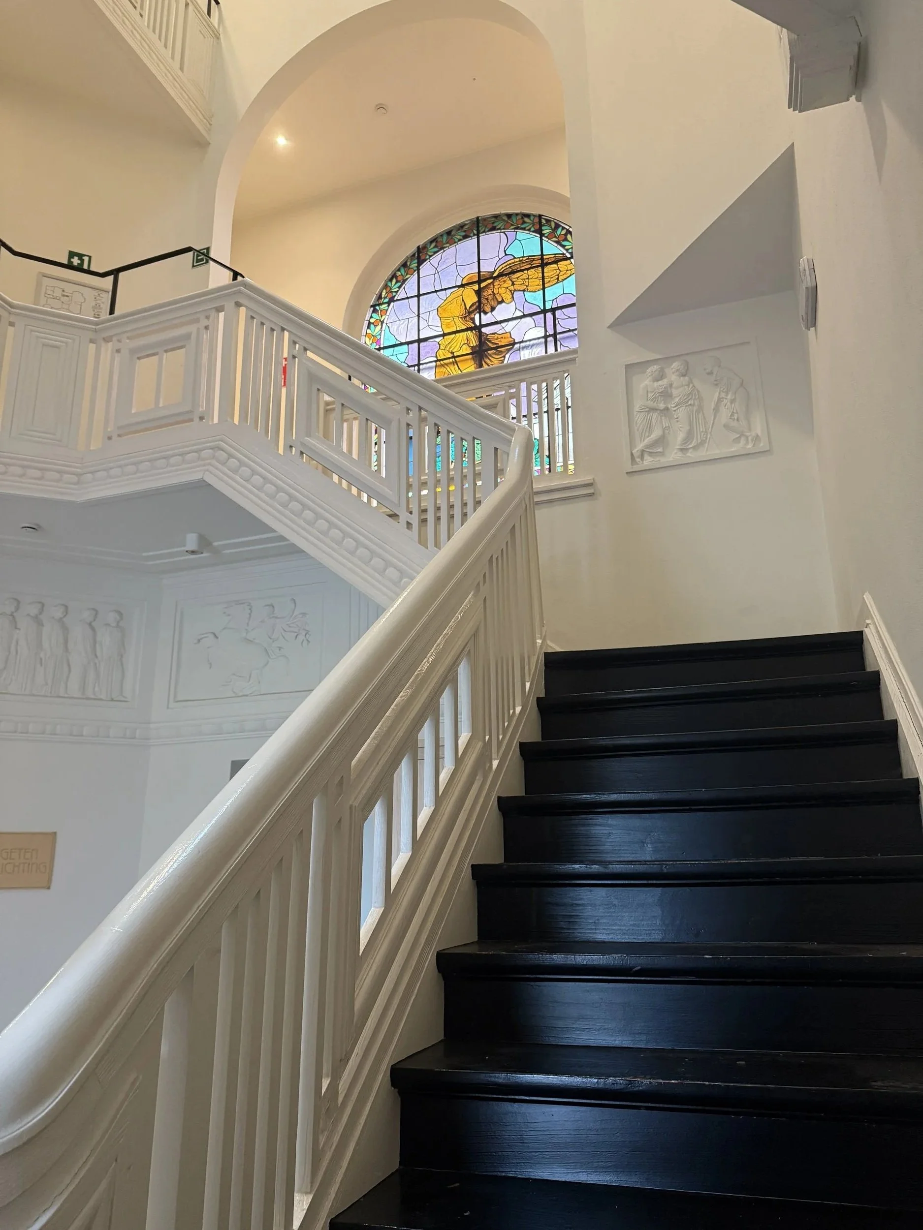Mooie binnentrap met zwarte houten treden en witte leuning in Villa Wybo te Zulte, met een gekleurd glas-in-lood raam op de achtergrond.