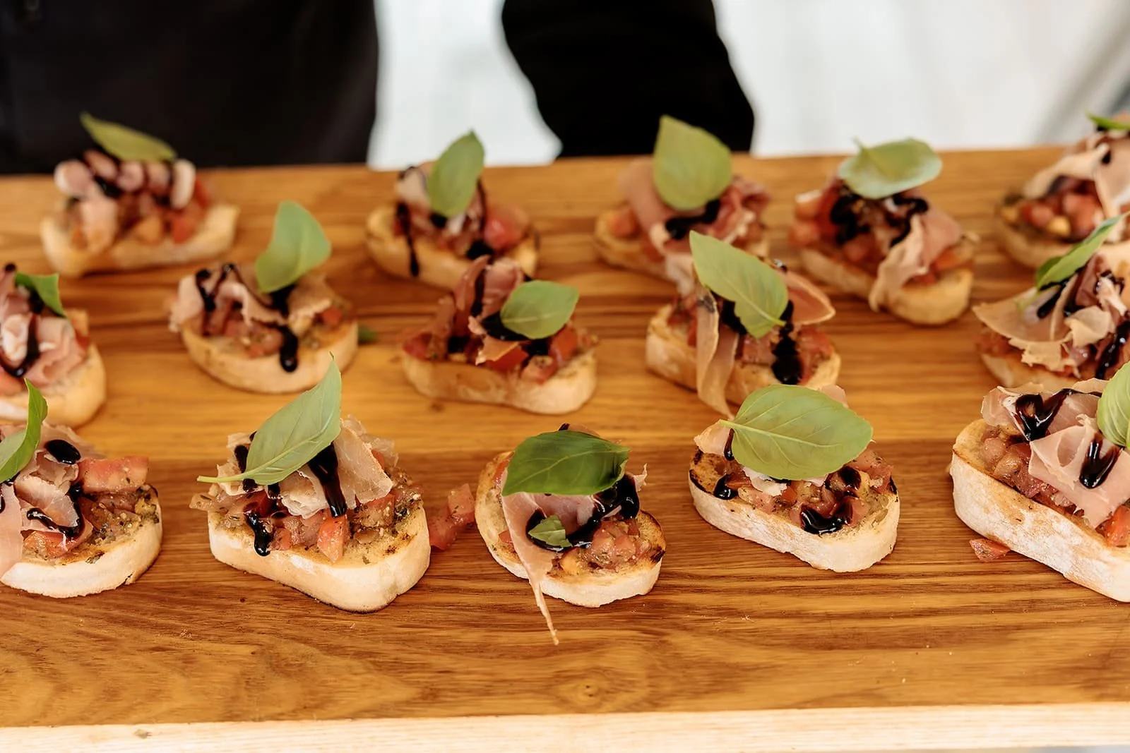 Hapjes met bruscetta en Parmaham, tomatensalsa met pesto, basilicum en balsamico dressing op een houten serveerschaal.