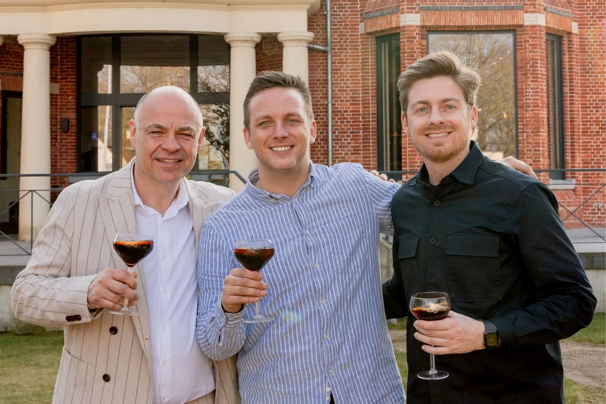 Drie mannen glimlachend met glazen rode wijn voor een groot, rode bakstenen gebouw met witte zuilen en grote ramen.