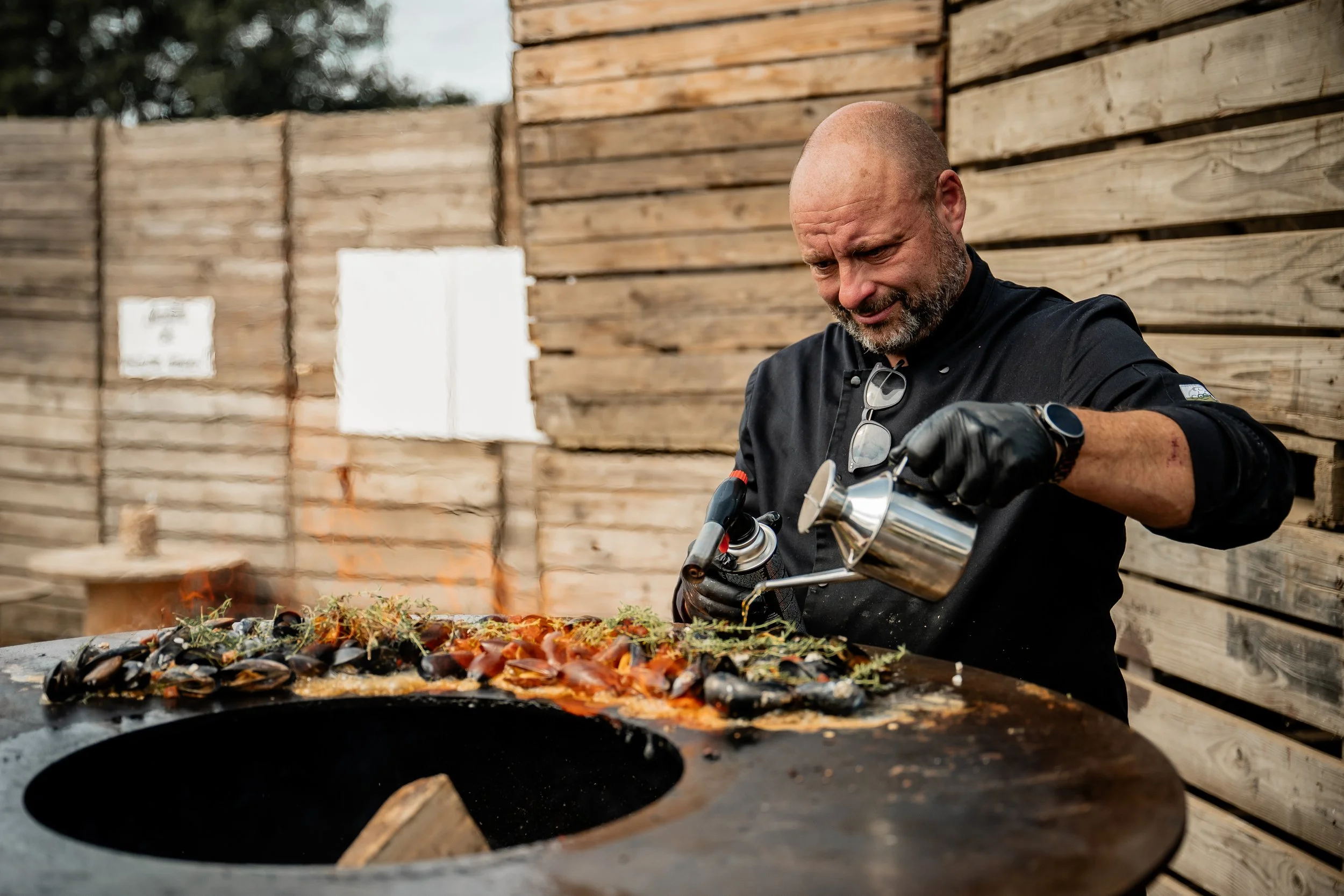 Man die op een open houtvuur mosselen bereidt, met een houten muur in de achtergrond.