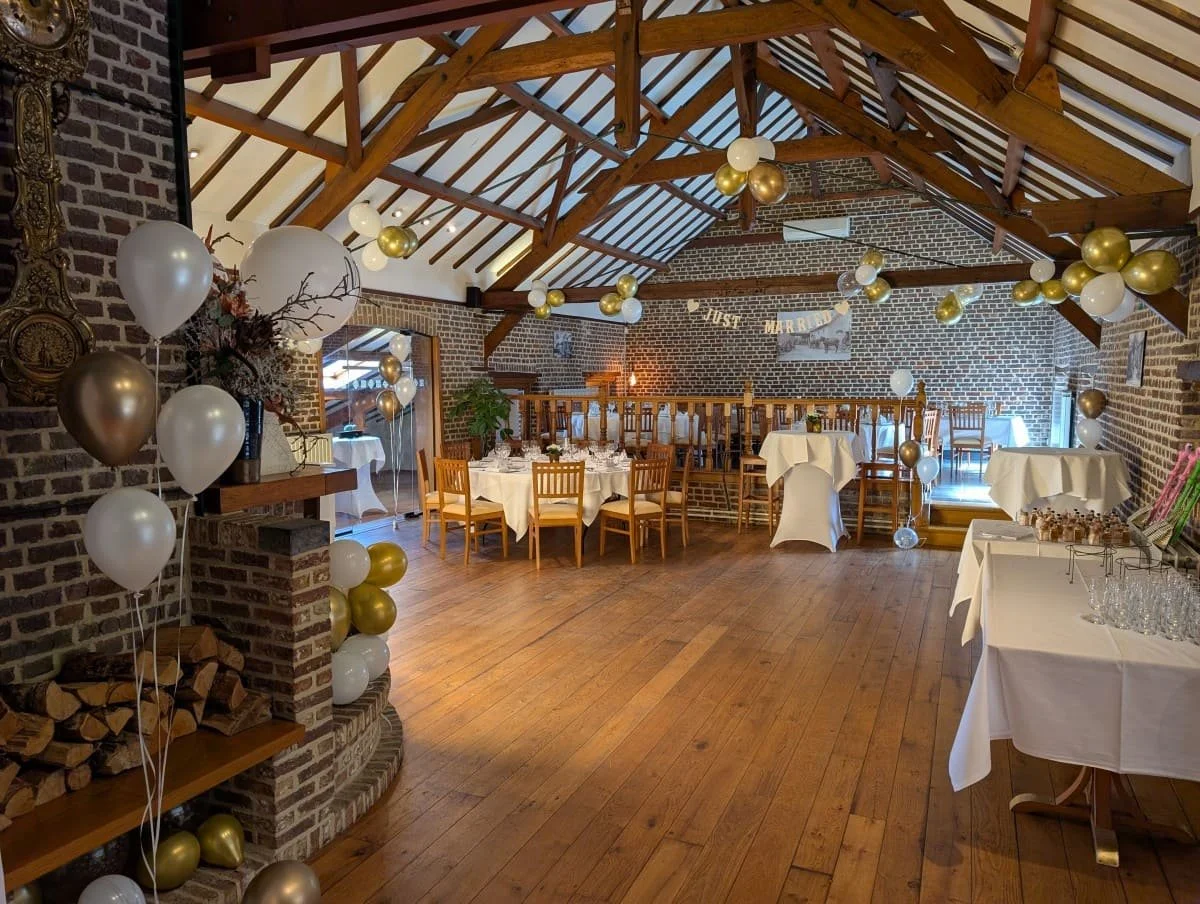 Versierde feestzaal met witte en gouden ballonnen, ronde tafels met witte tafelkleden, houten vloer en brick muren, decoraties en lichtjes, klaar voor een feest of bruiloft.