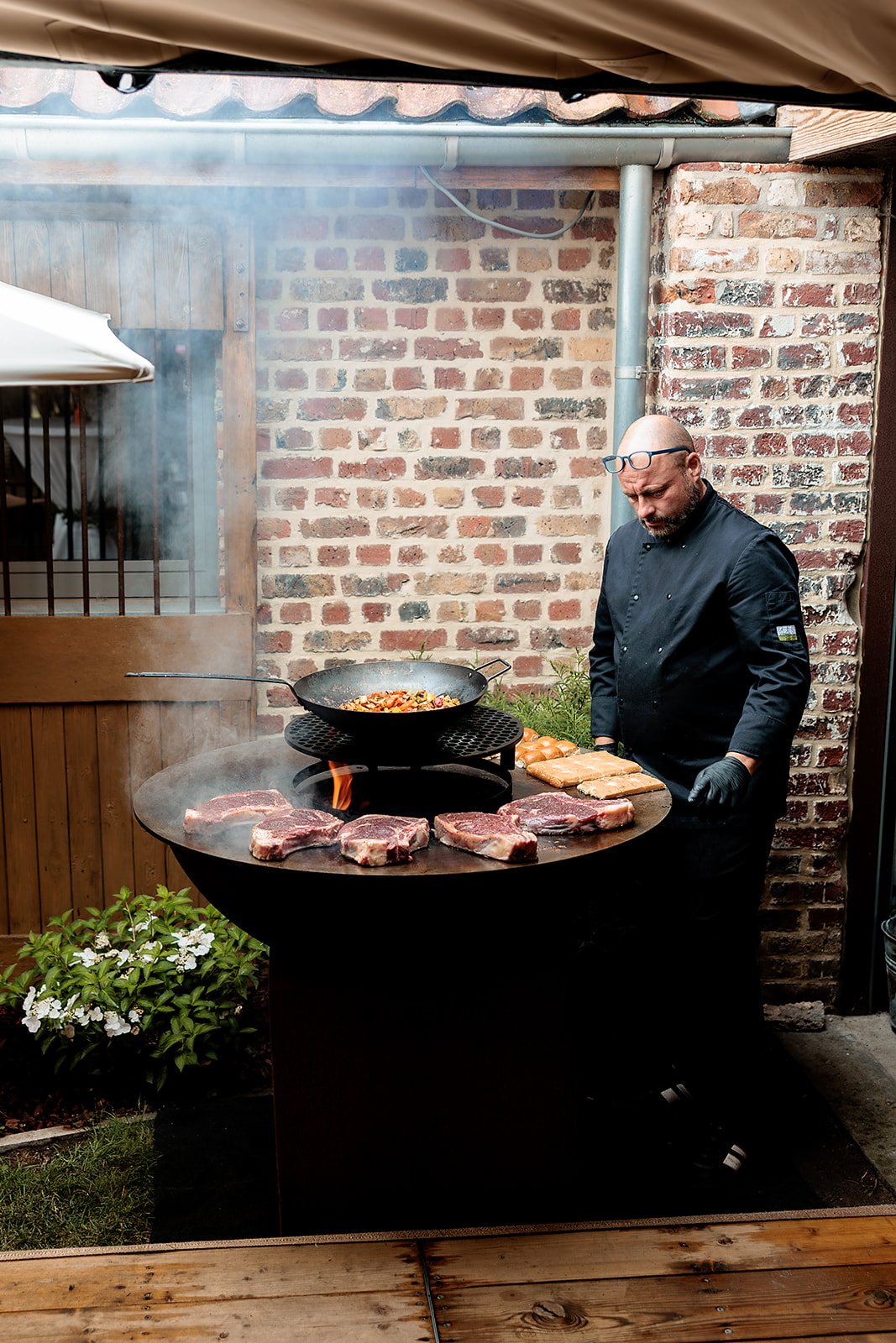 Een kok bereidt vlees op een barbecue buiten met een zandstenen muur op de achtergrond.