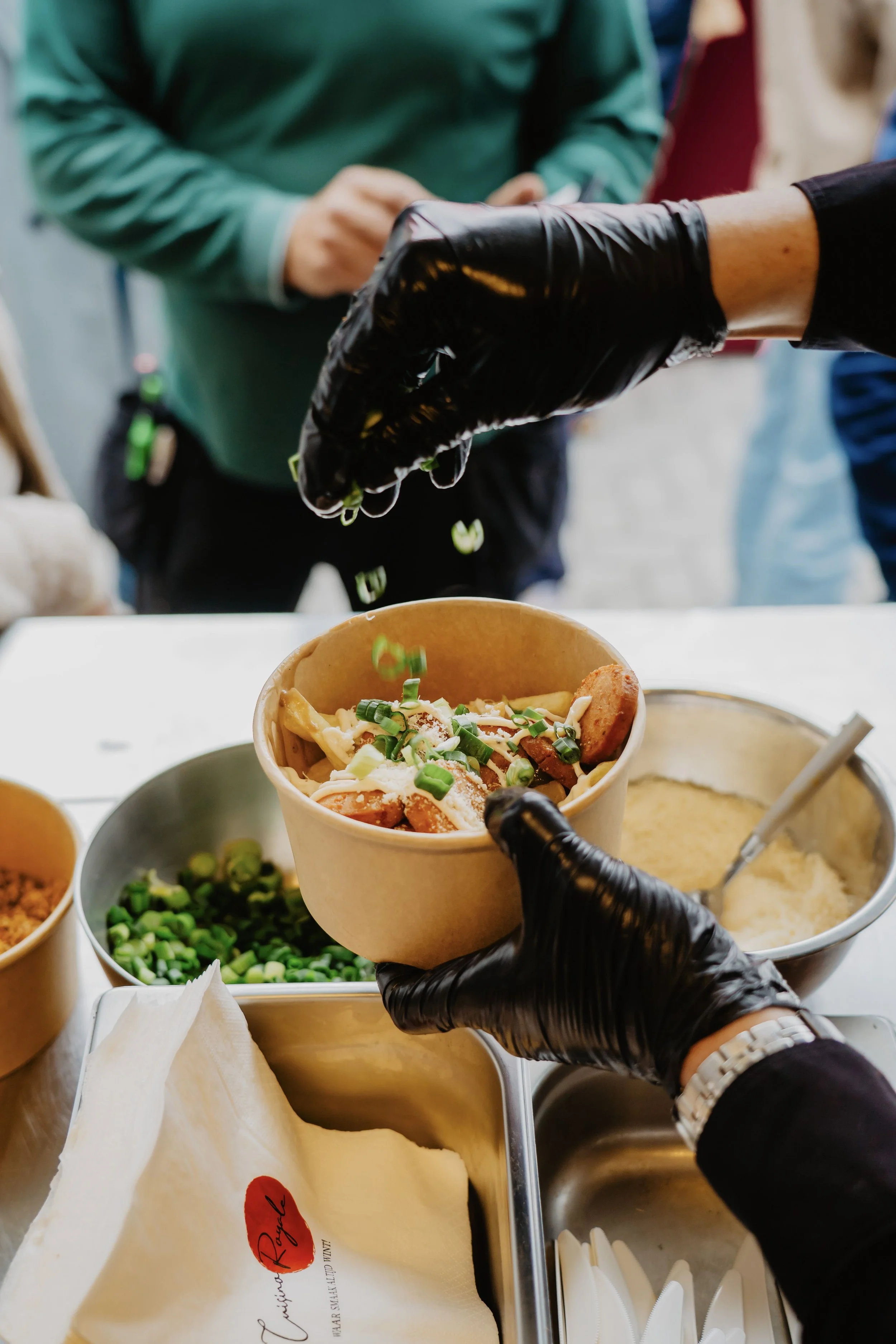 Een persoon in een zwart handschoen die een kom met friet en groenten vasthoudt, terwijl een andere persoon in een zwart handschoen zout over de friet strooit. Op de tafel staan verschillende schaaltjes met groenten en saus.