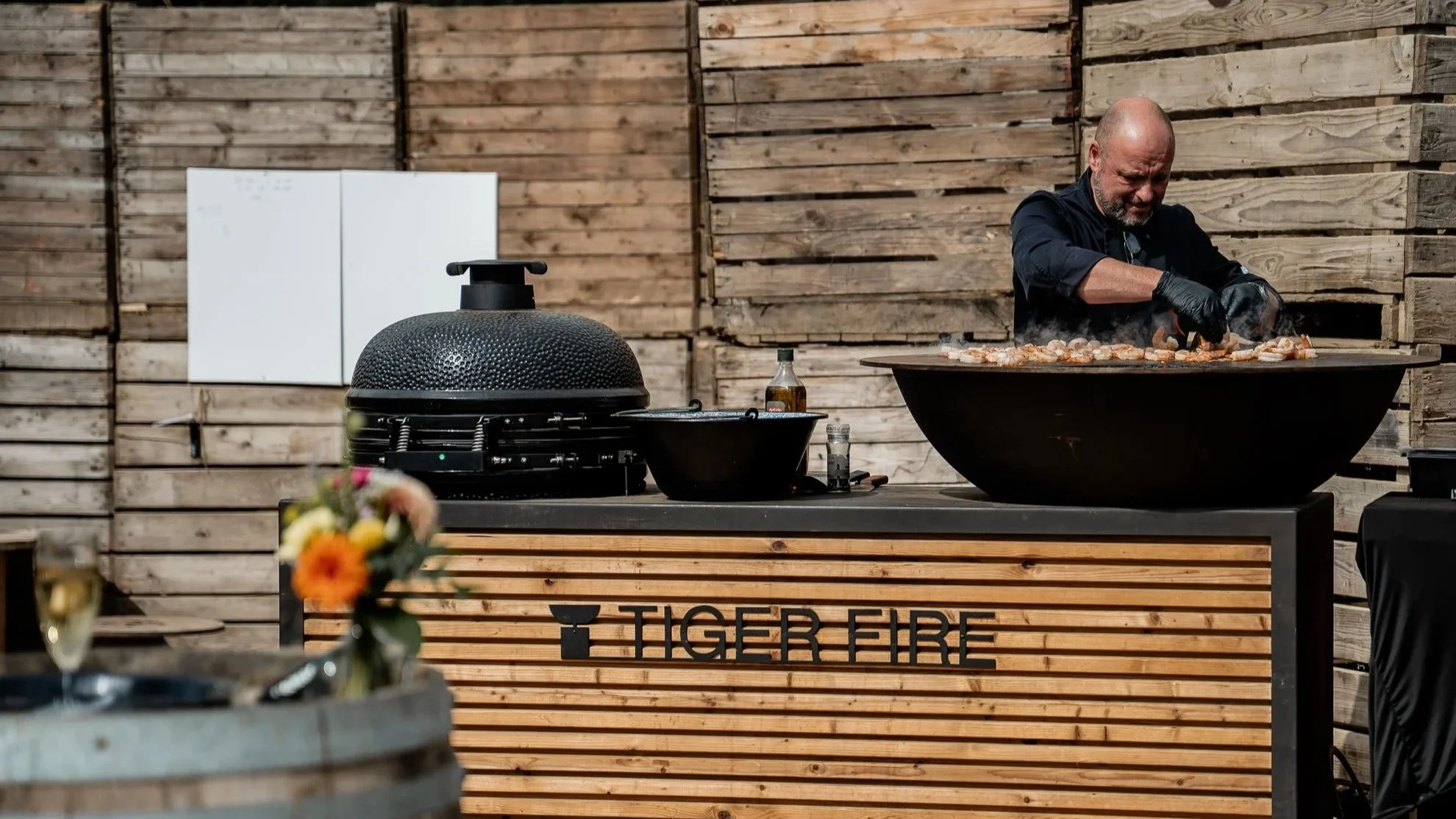 Een mannelijke chef bereidt voedsel op een grote zwarte barbecue met de tekst 'TIGER FIRE' op het houtachtige aanrecht. Op de achtergrond een houten muur.