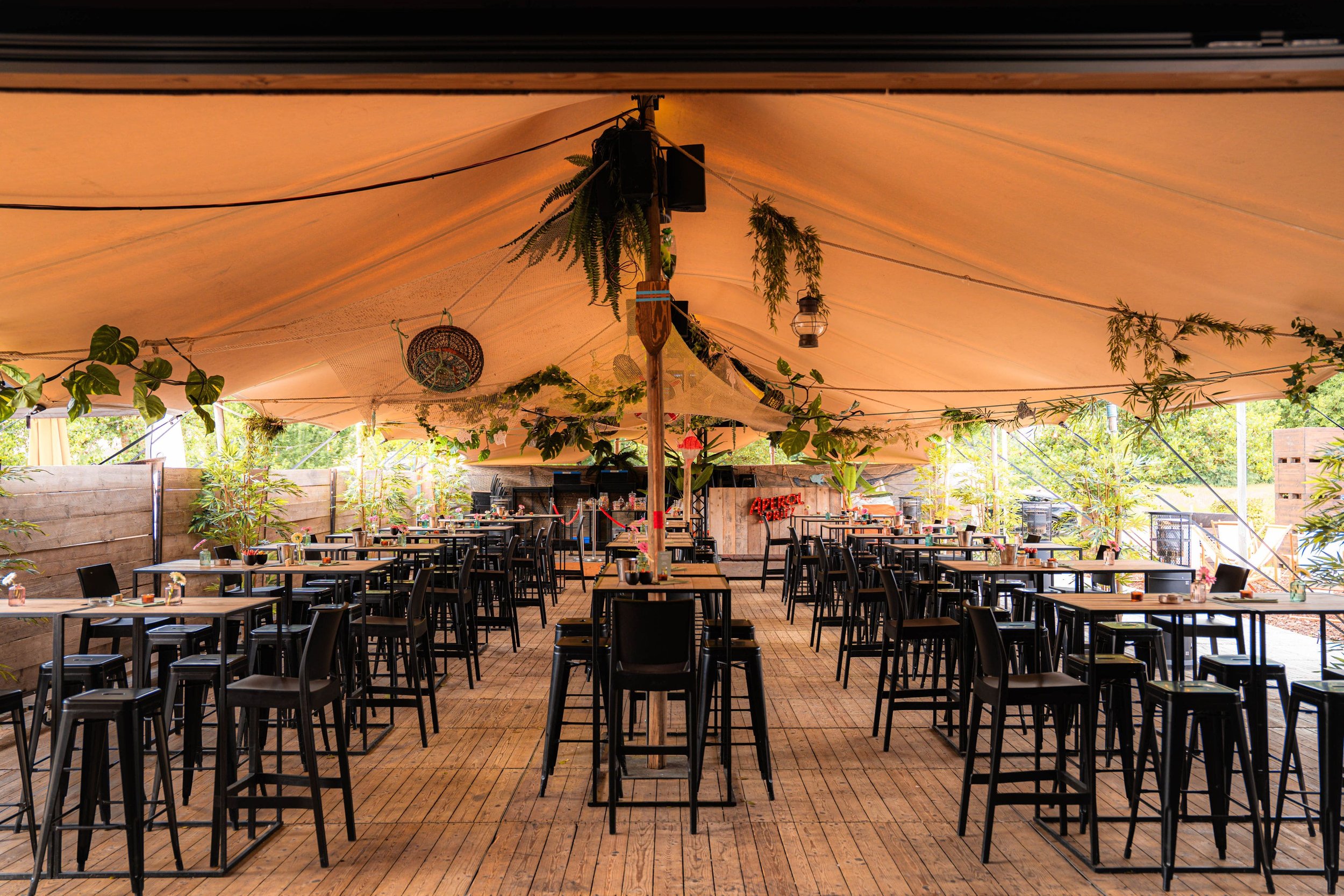 Een tuinfeest tent met hoge plafonds en houten vloer, ingericht met zwarte statafels en stoelen, versierd met groen en lichtslingers.