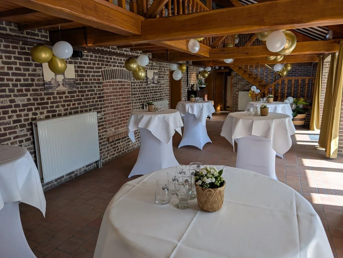 Binnenruimte met ronde tafels met witte tafelrokken en een bloemstuk, versierd met gouden en witte ballonnen voor een feest of bijeenkomst.