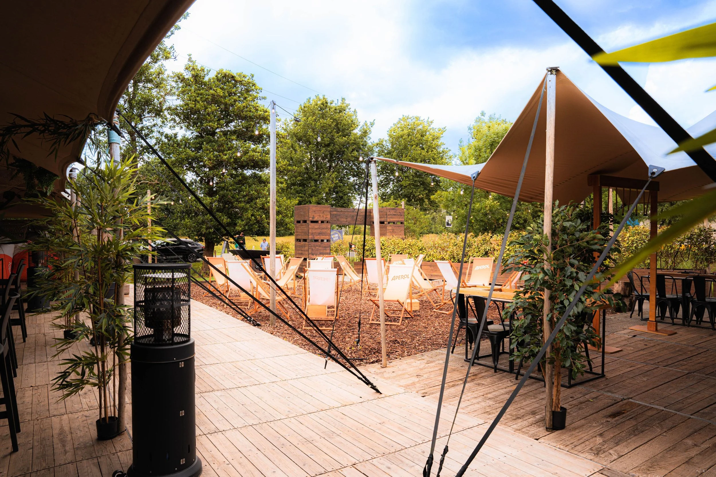 Een buitenfaciliteit met houten terrassen, zonneschermen, planten, en lege ligstoelen en tafels onder een blauwe lucht.