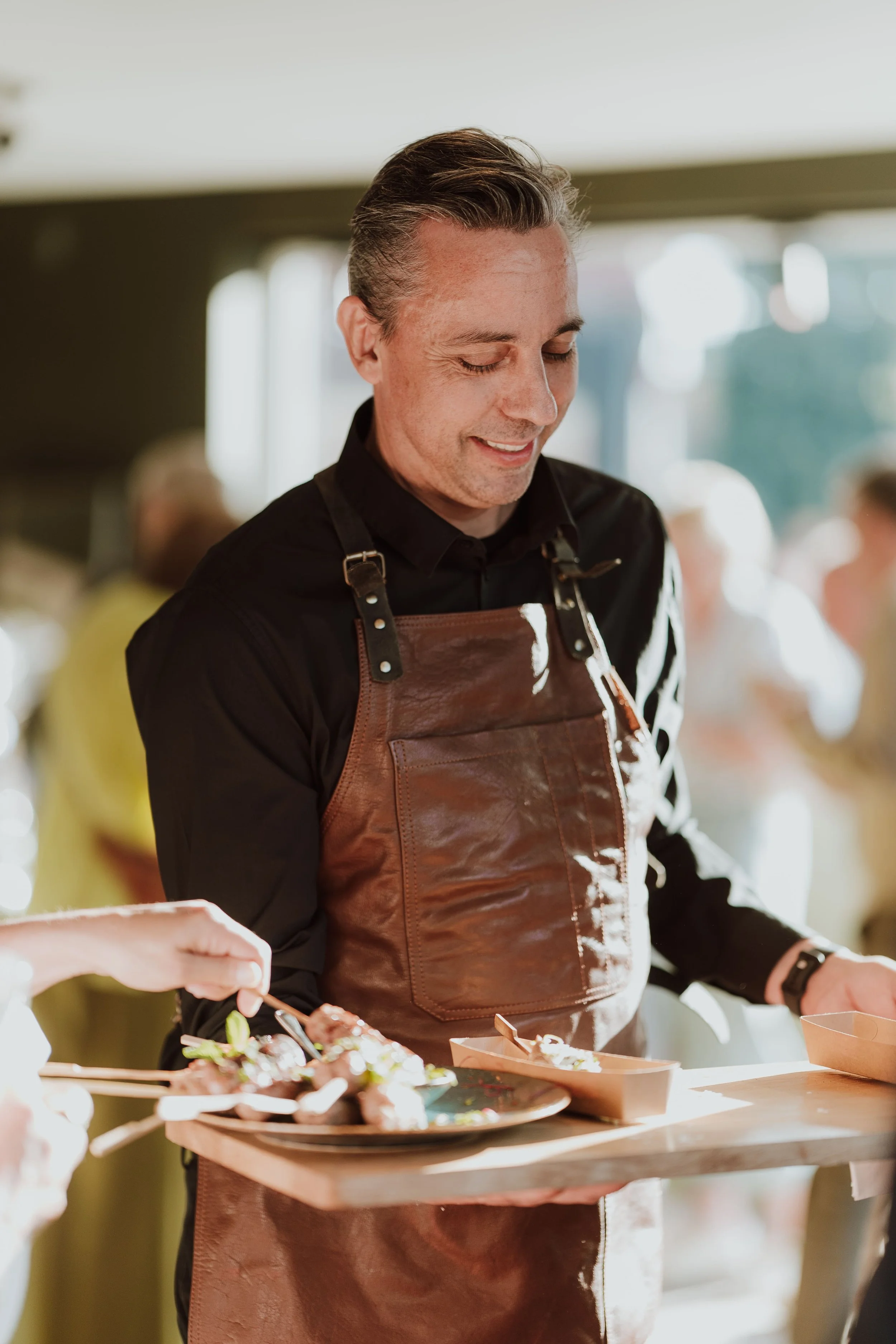 Man in een zwarte overhemd en bruin leren schort serveert voedsel op een dienblad, glimlachend in een lichte, gezellige setting.