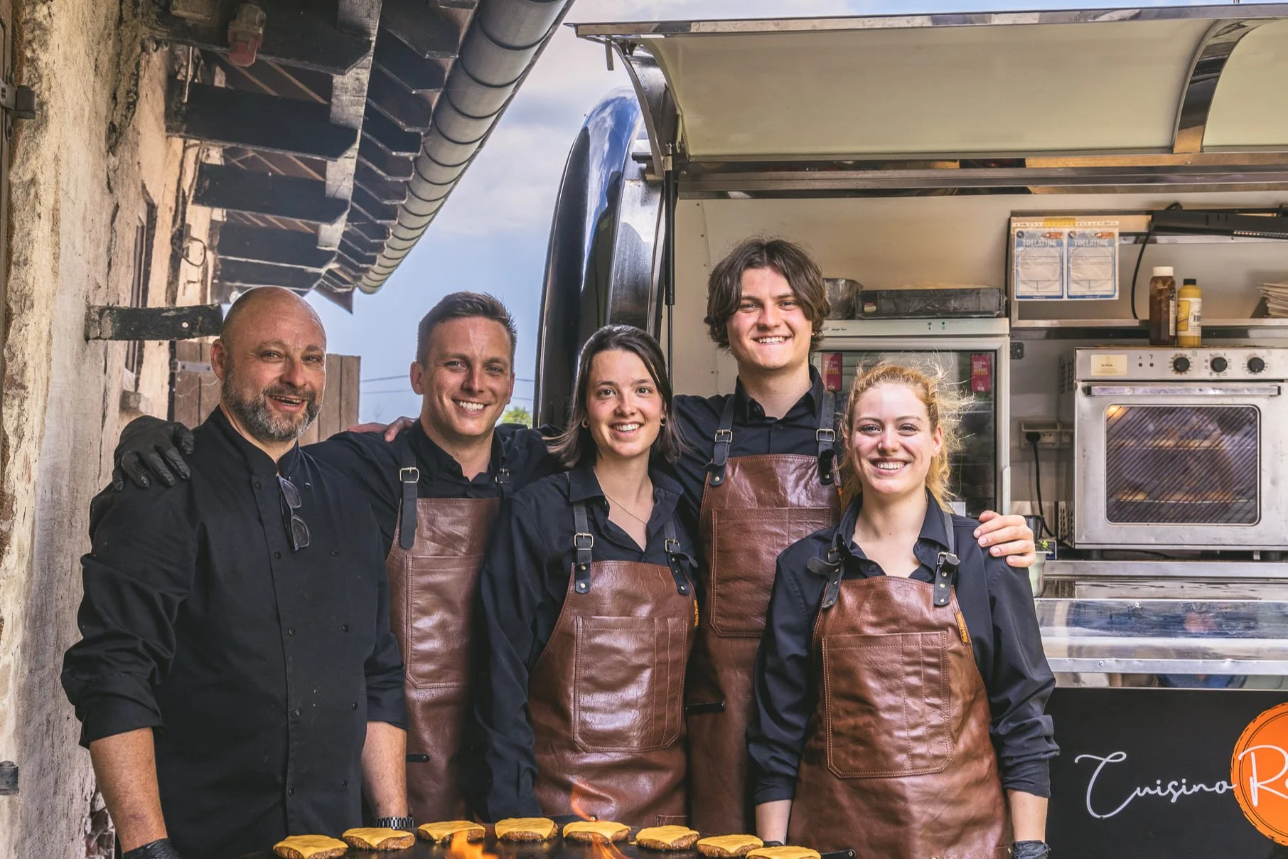 Vijf mensen in zwarte kleren en bruin leren schorten poseert samen voor een foodtruck, met koekjes op de tafel voor hen.