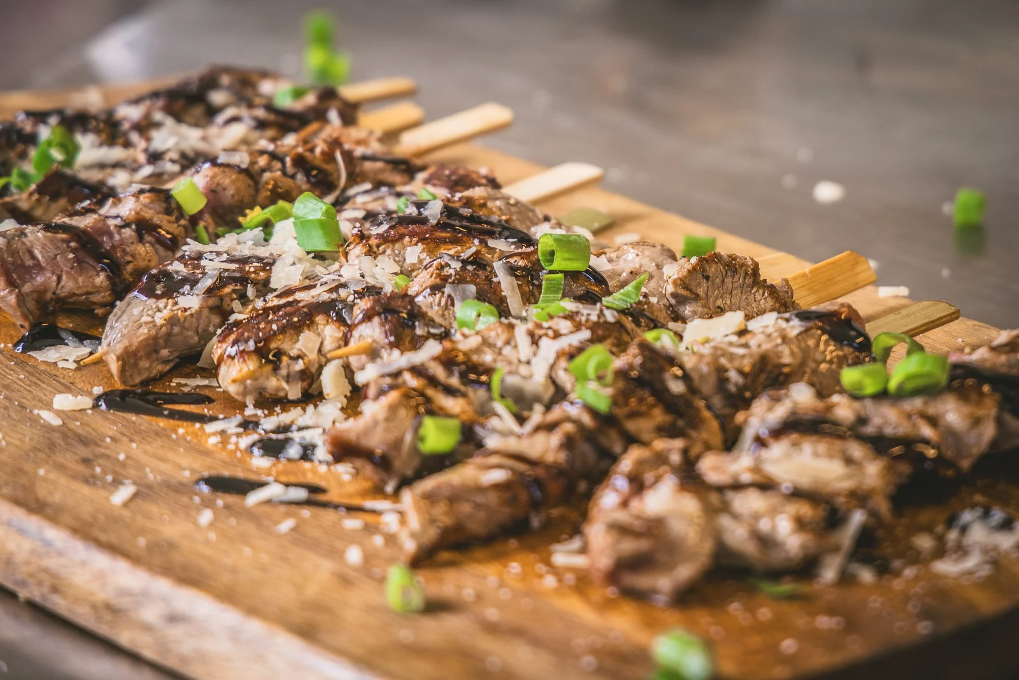 Stukjes rundsvlees op een spiesje met groene uien, geschaafde Parmesaanse kaas en balsamico saus op een houten snijplank.