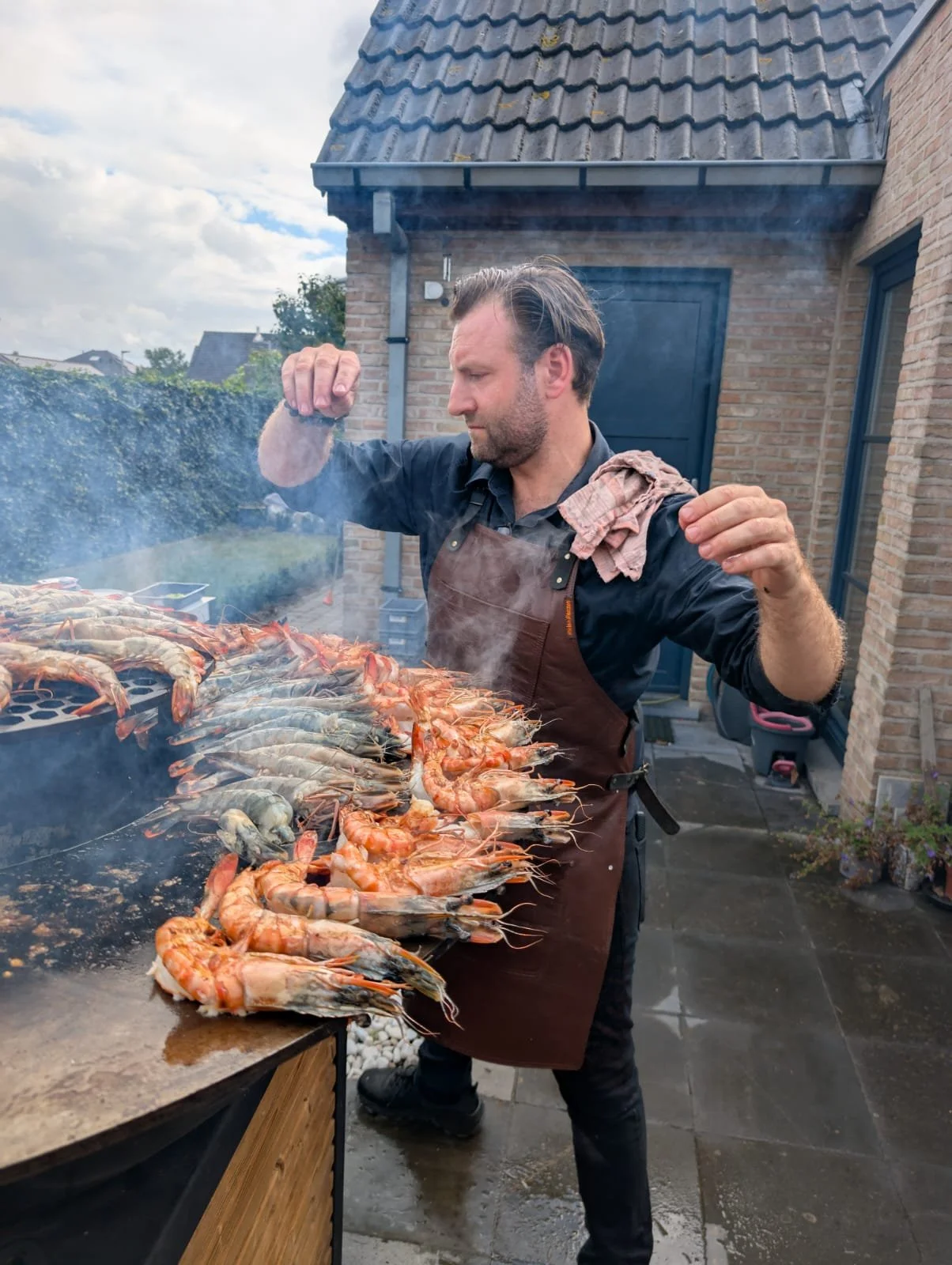 Man die gamba's grilt op een buitengrill, met een achtergrond van een huis en een tuin.