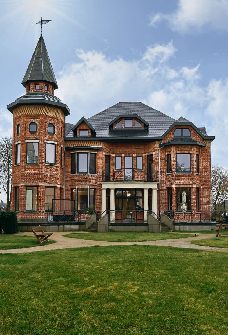 Een groot, stenen huis met een toren en veel ramen, gelegen op een groen grasveld met wandelpaden en bankjes.