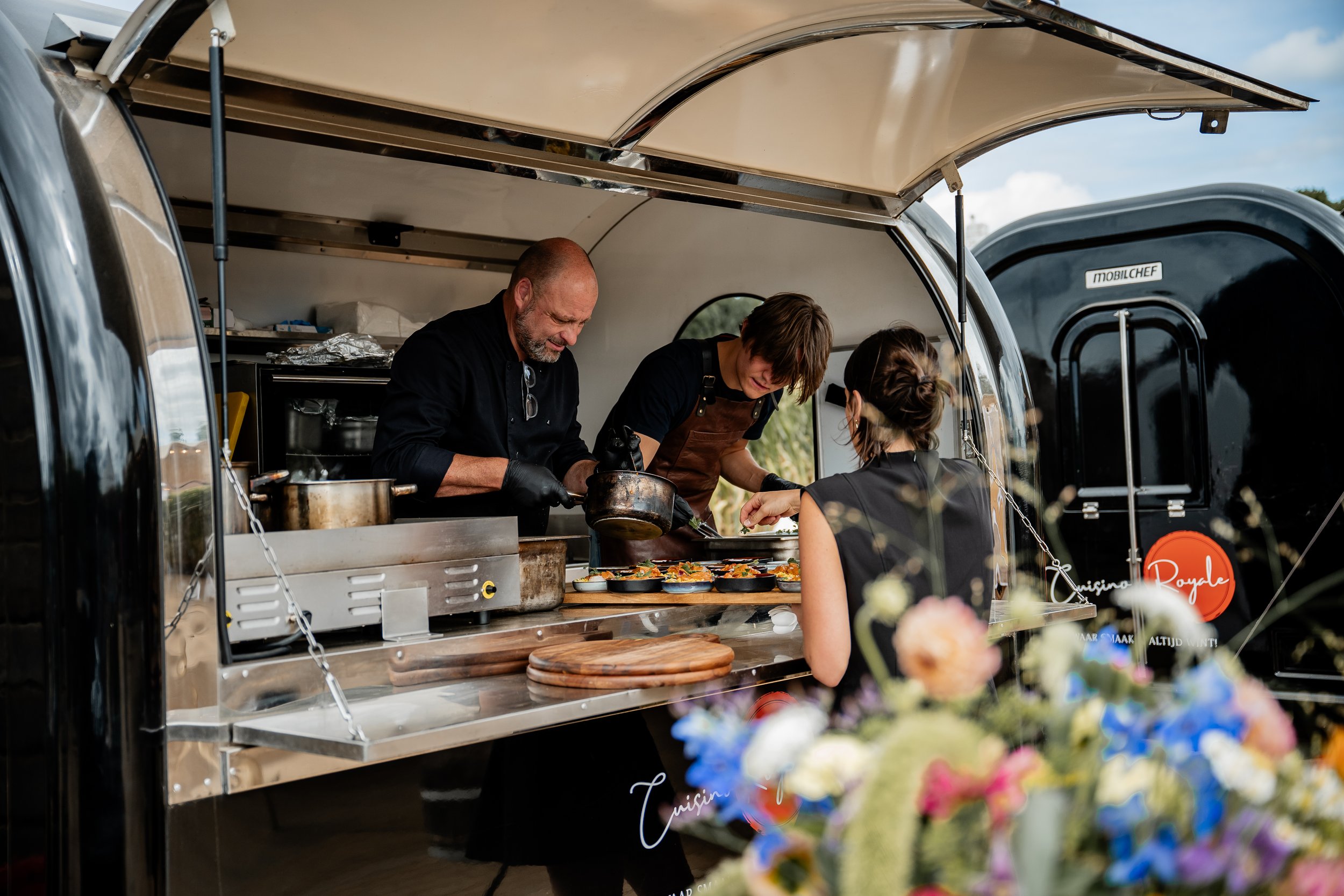 Kokende chef en souschef serveren gerechten aan een klant vanuit een foodtruck met bloemen op de voorgrond.