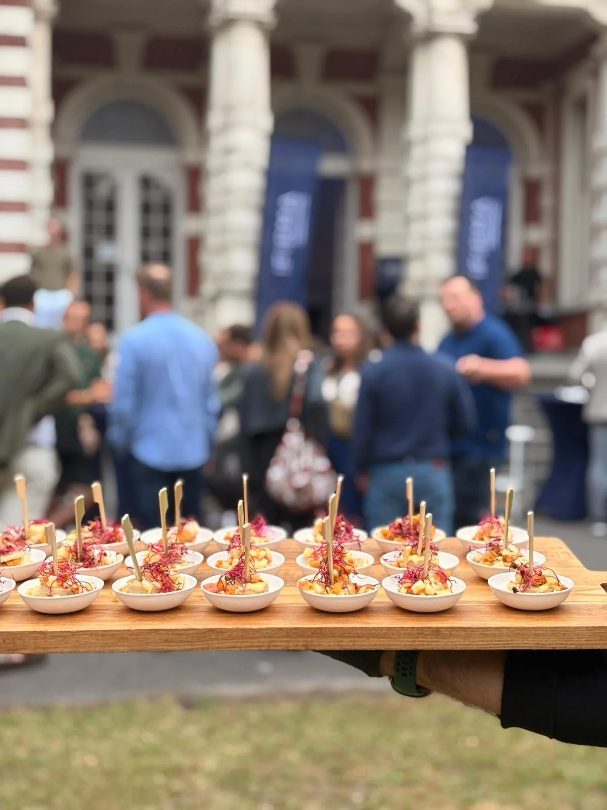 Een hand die een plank met kleine hapjes vasthoudt voor een groep mensen die buiten voor een historisch gebouw staan.