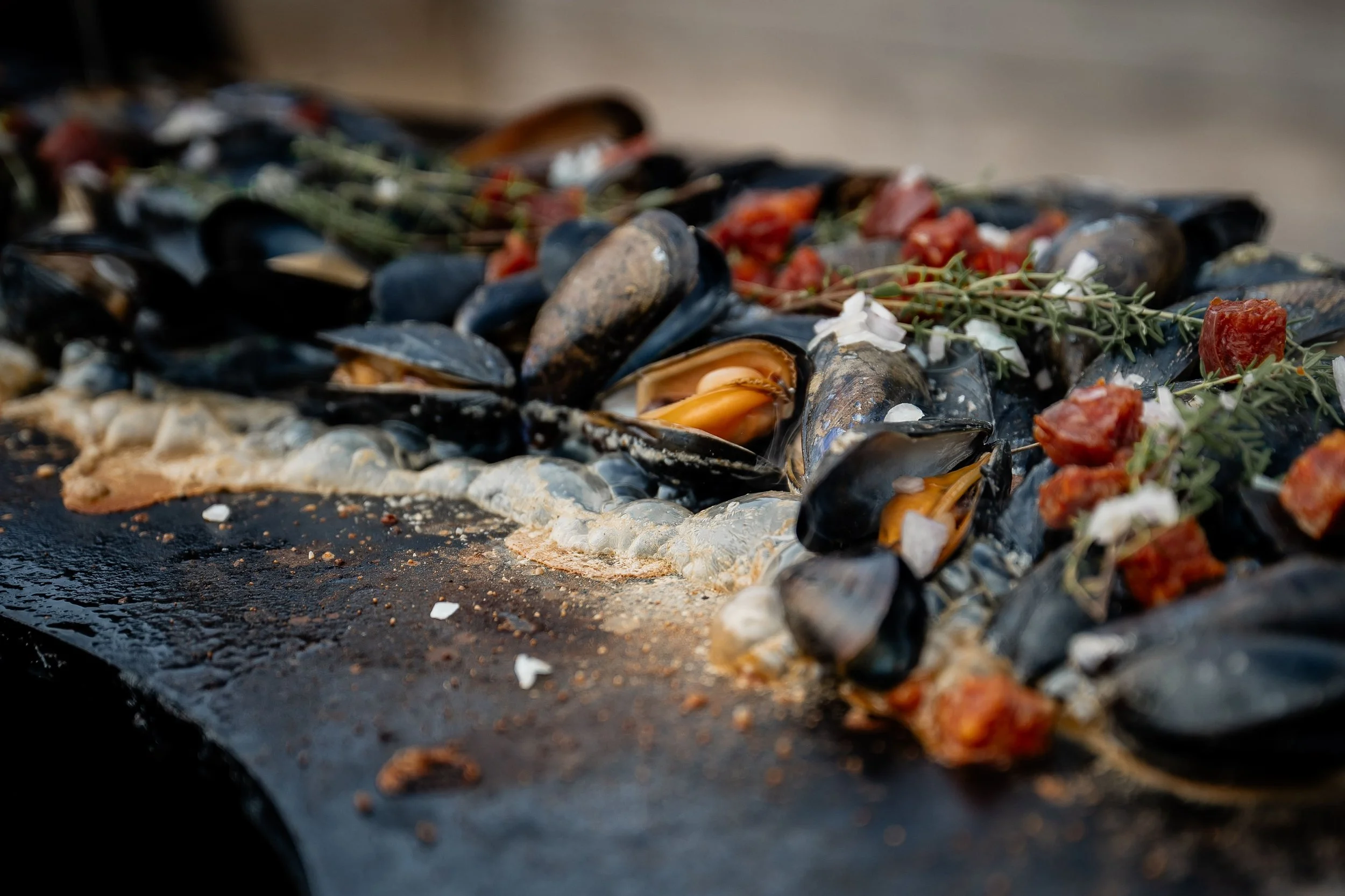 Close-up van een gegrilde mosseltjes met garnituur van chorizo, ui en tijm, geserveerd op een zwarte bakplaat.