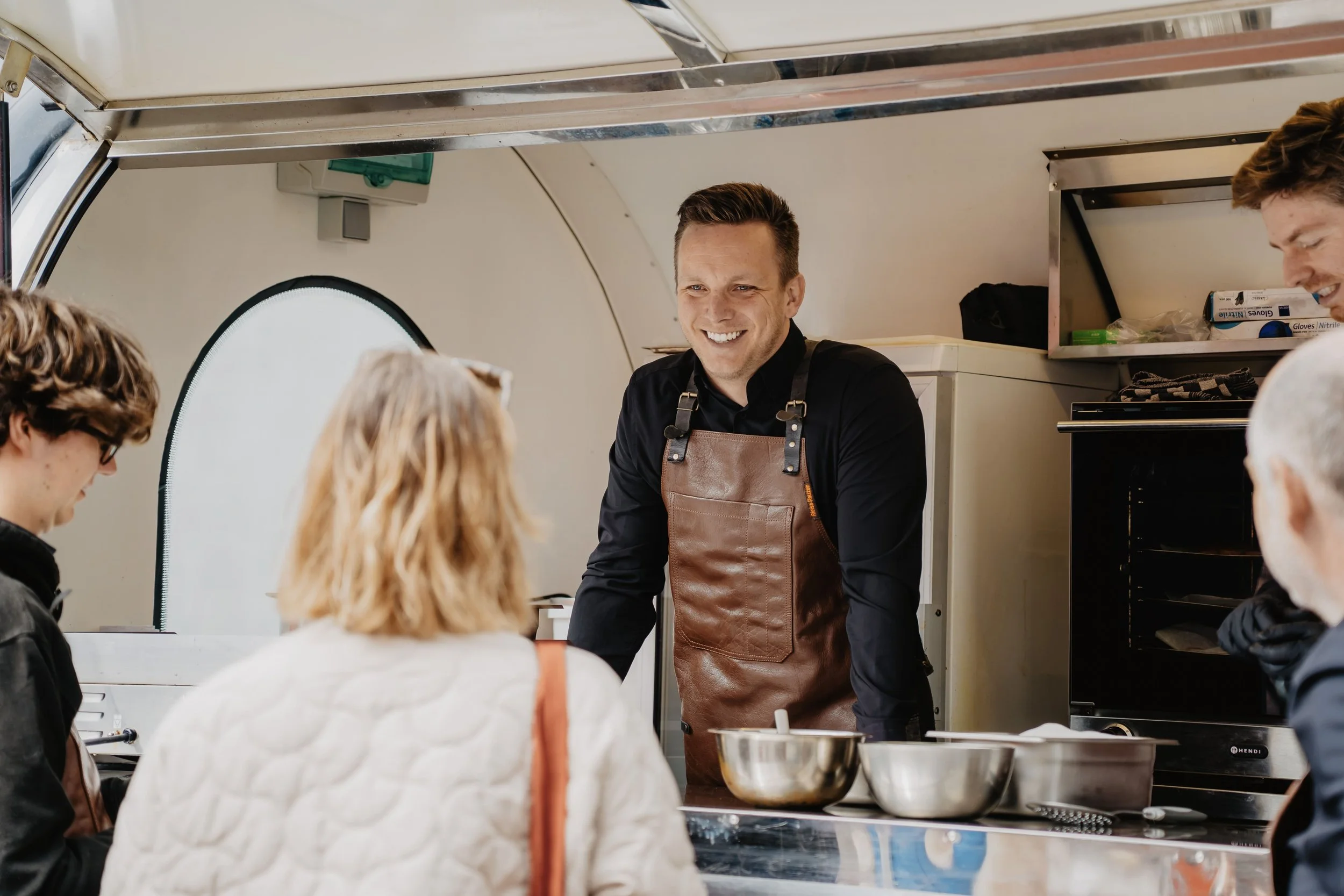 Man in een bruin leren schort glimlacht terwijl hij met klanten in een foodtruck praat.