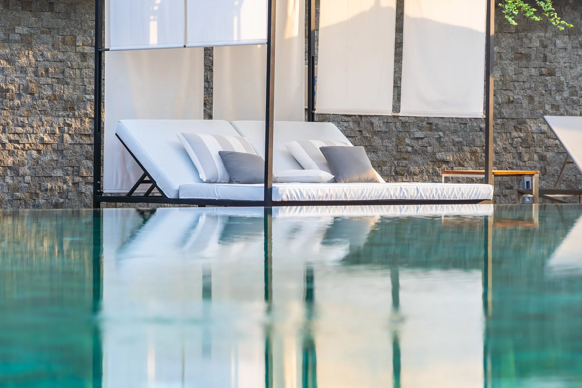 Poolside lounge chair with white mattress and pillows, partially shaded by a canopy, next to a brick wall.