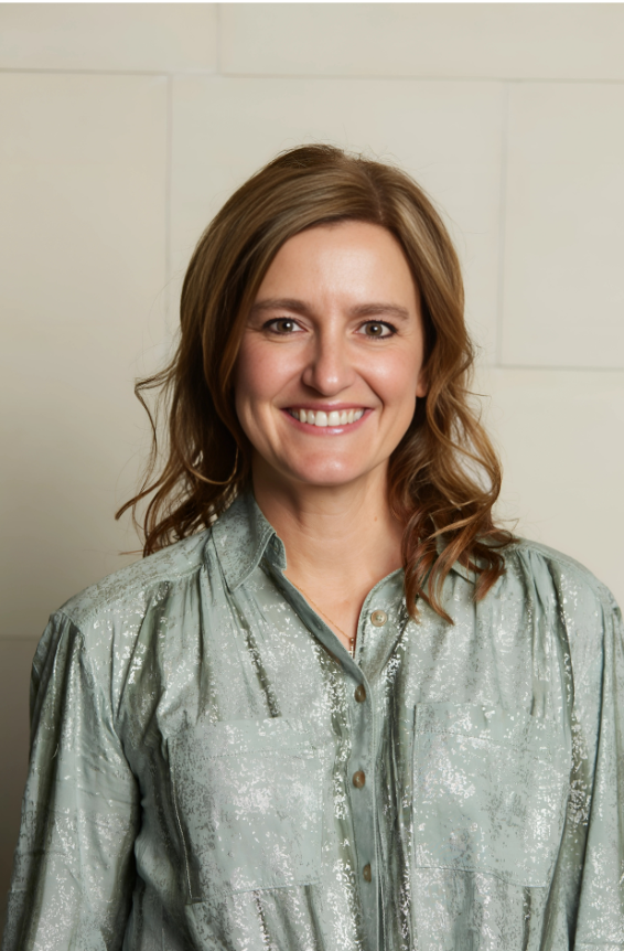 A woman with shoulder-length light brown hair, smiling, wearing a shiny silver-green button-up blouse, standing in front of a light-colored tiled wall.