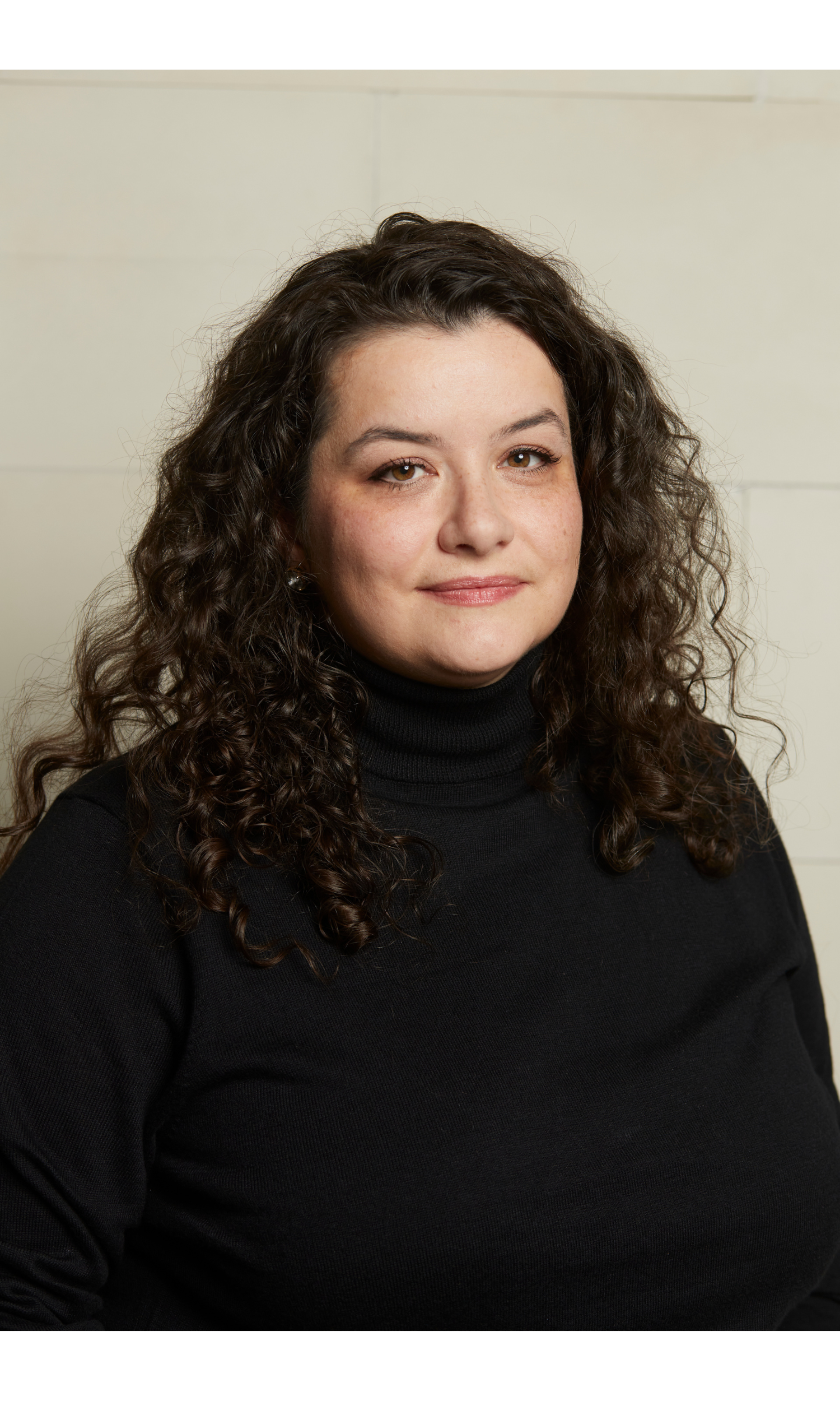A woman with long, curly dark hair and fair skin posing in front of a beige wall, wearing a black turtleneck sweater, looking at the camera with a slight smile.