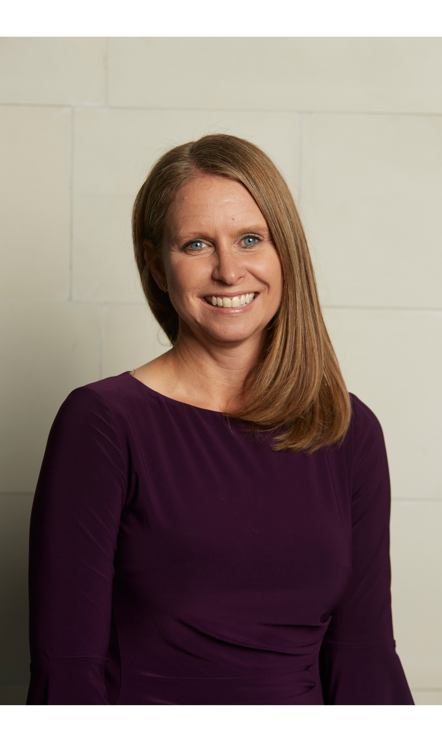 Headshot of a woman with long light brown hair, wearing a dark purple top, smiling, standing against a plain off-white wall.