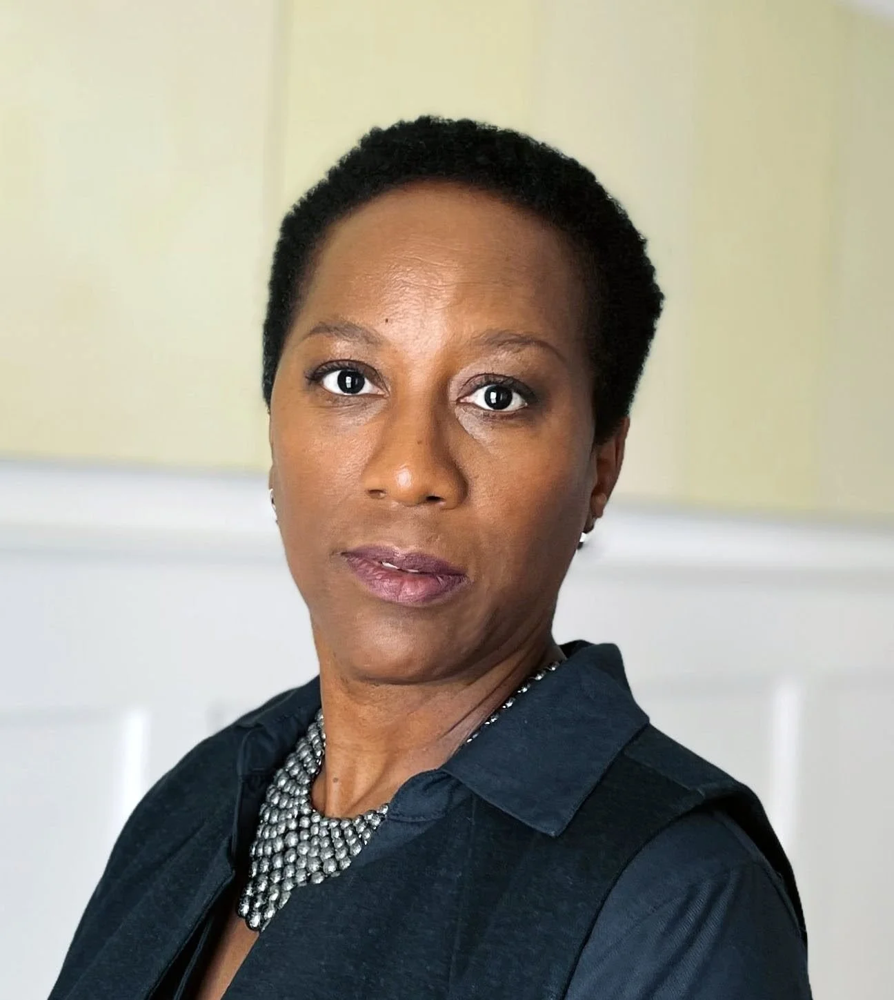 A woman with short black hair, wearing a black top and a silver beaded necklace, stands indoors with neutral-colored walls in the background.