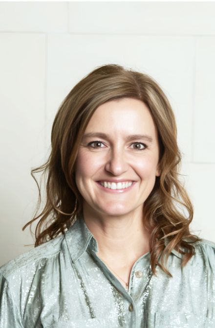 A woman with light brown wavy hair smiling, wearing a light-colored shiny blouse, posing against a plain white wall.