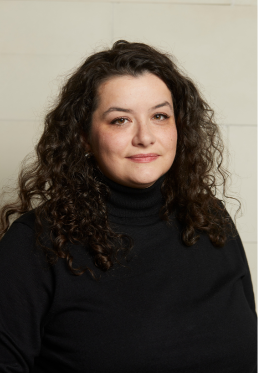 A woman with curly brown hair wearing a black turtleneck, standing against a light-colored wall.