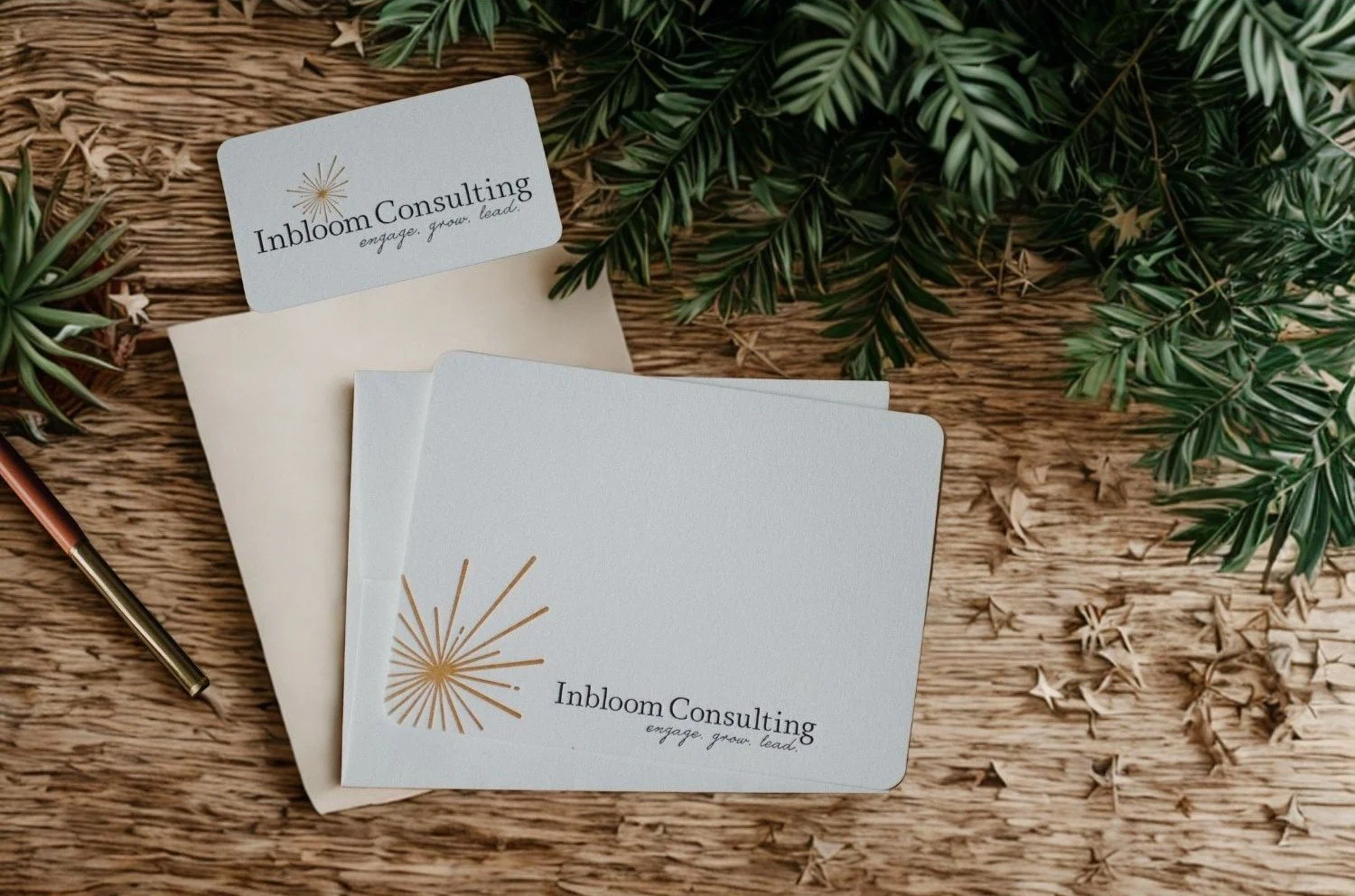 Business cards for Inbloom Consulting placed on a rustic wooden surface with green foliage, a pen, and an envelope.