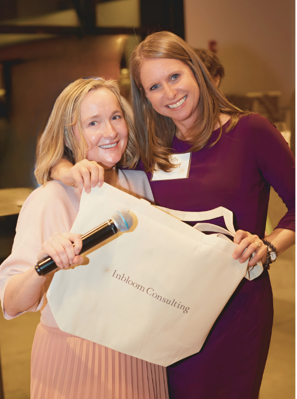Two women smiling at an event, one holding a microphone, the other holding a bag with the logo 'Inbloom Consulting'.