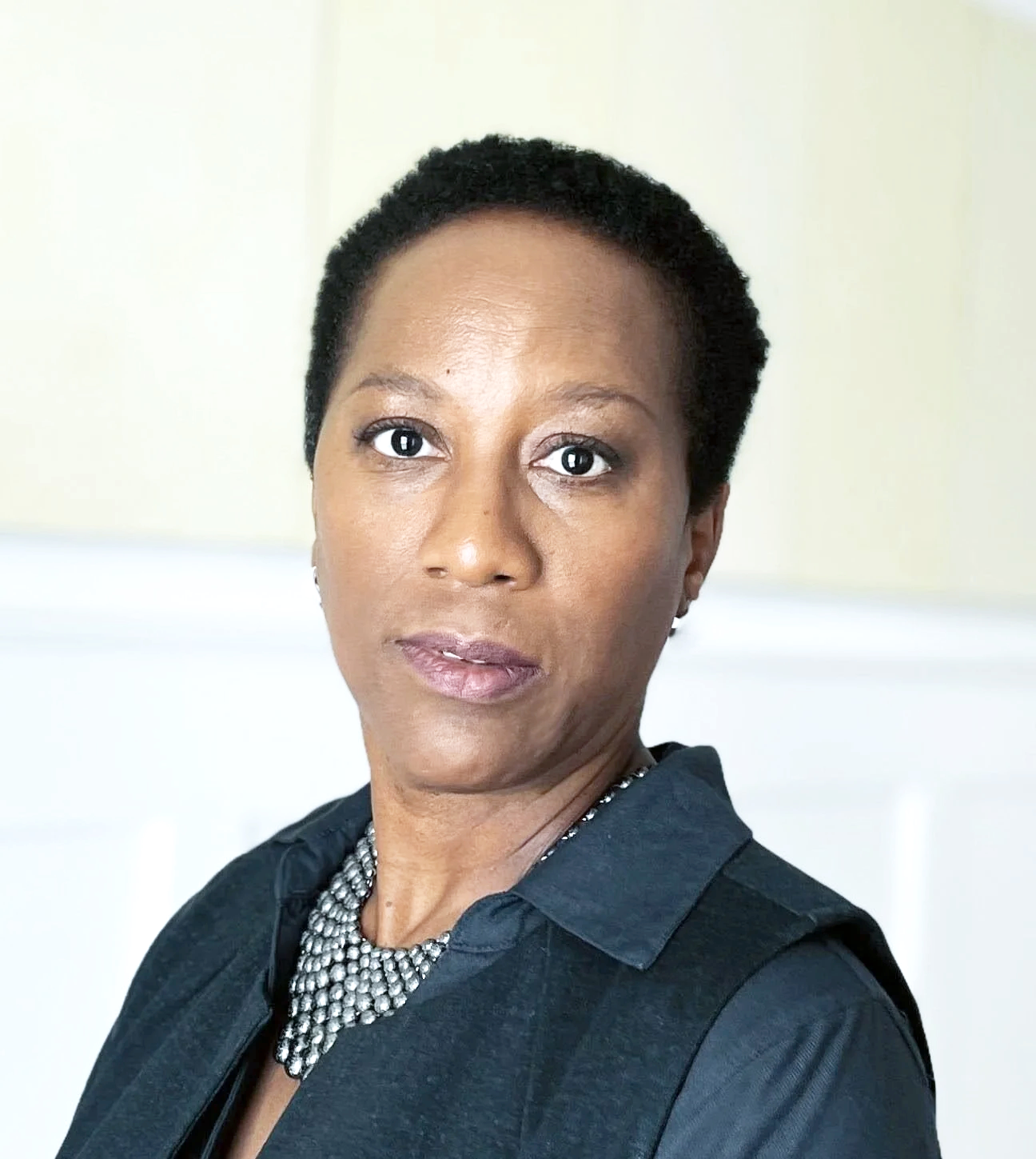 A portrait of an African American woman with short curly black hair, wearing a black top and a silver necklace, standing against a neutral background.