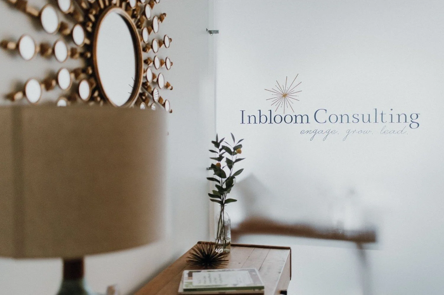 Interior of an office with a wall displaying the Inbloom Consulting logo and slogan, a small table with a vase holding a branch, and a decorative mirror on the wall.