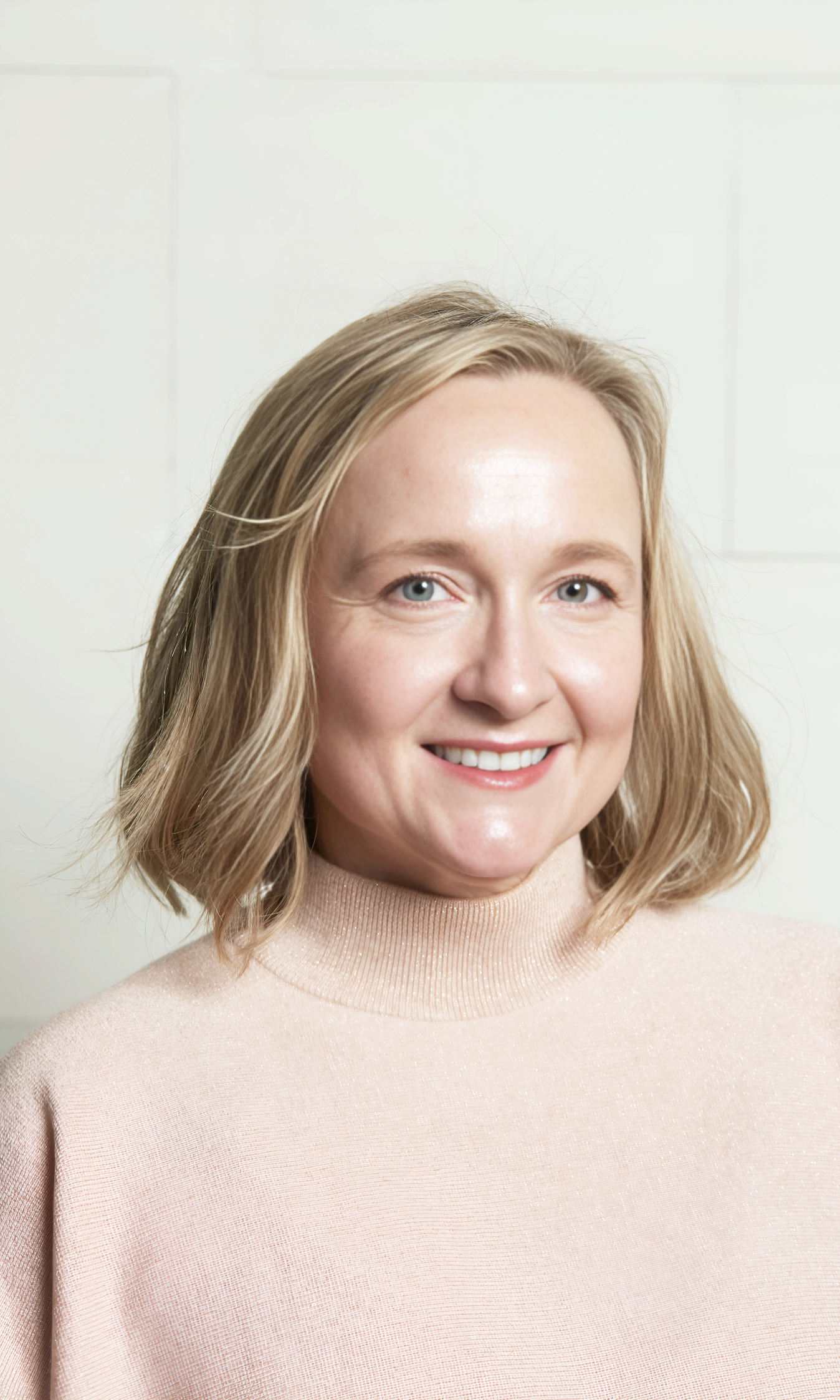 A woman with shoulder-length blonde hair, blue eyes, and fair skin smiling, wearing a light pink turtleneck sweater, standing in front of a light-colored wall.