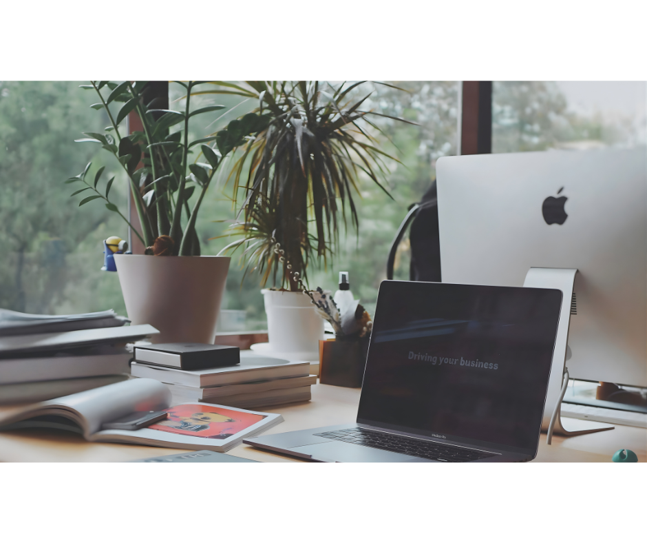 Office desk near a window with potted plants, notebooks, magazines, a laptop, and an iMac computer.