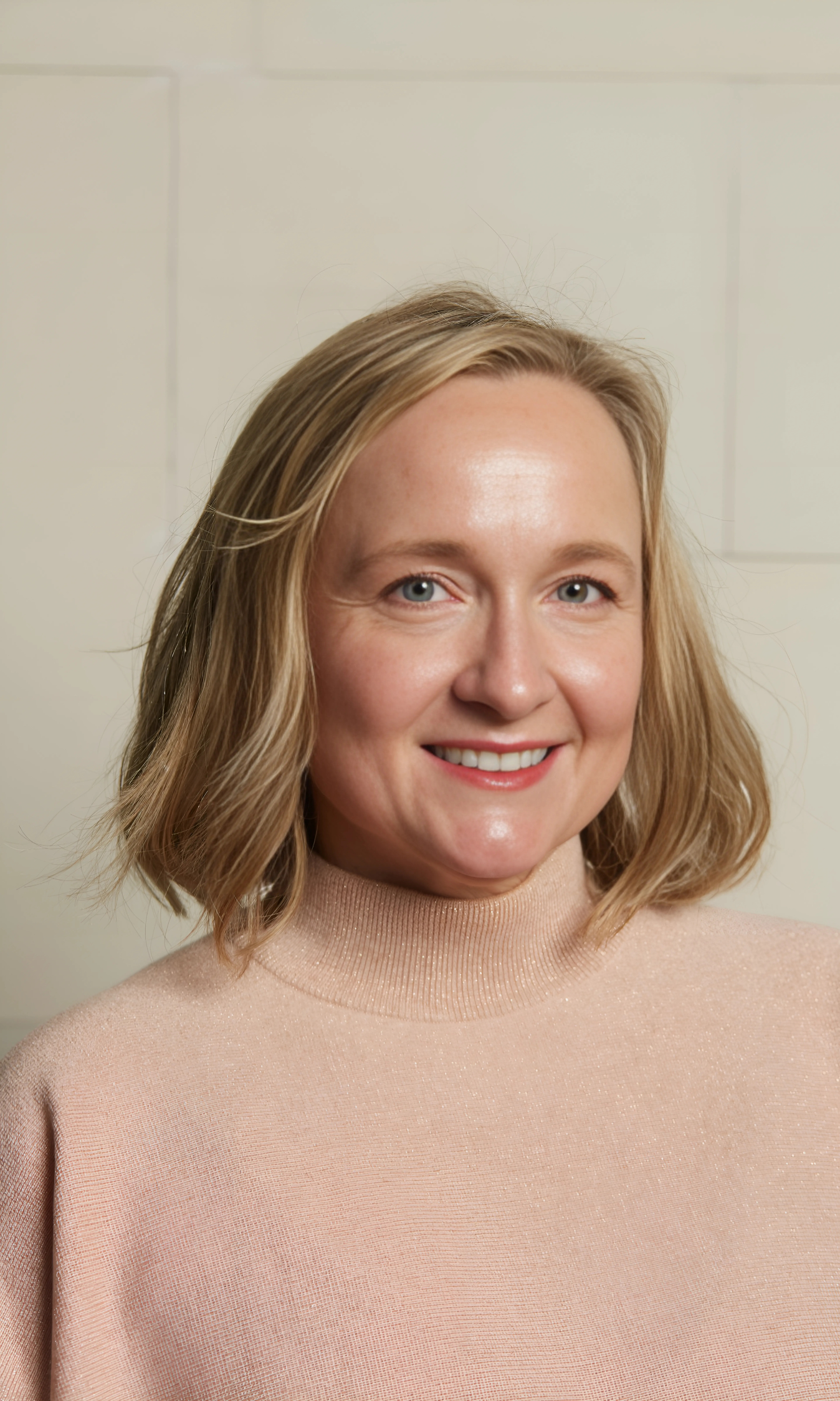 Close-up of a smiling woman with shoulder-length blonde hair, wearing a light pink sweater, standing in front of a plain white wall.