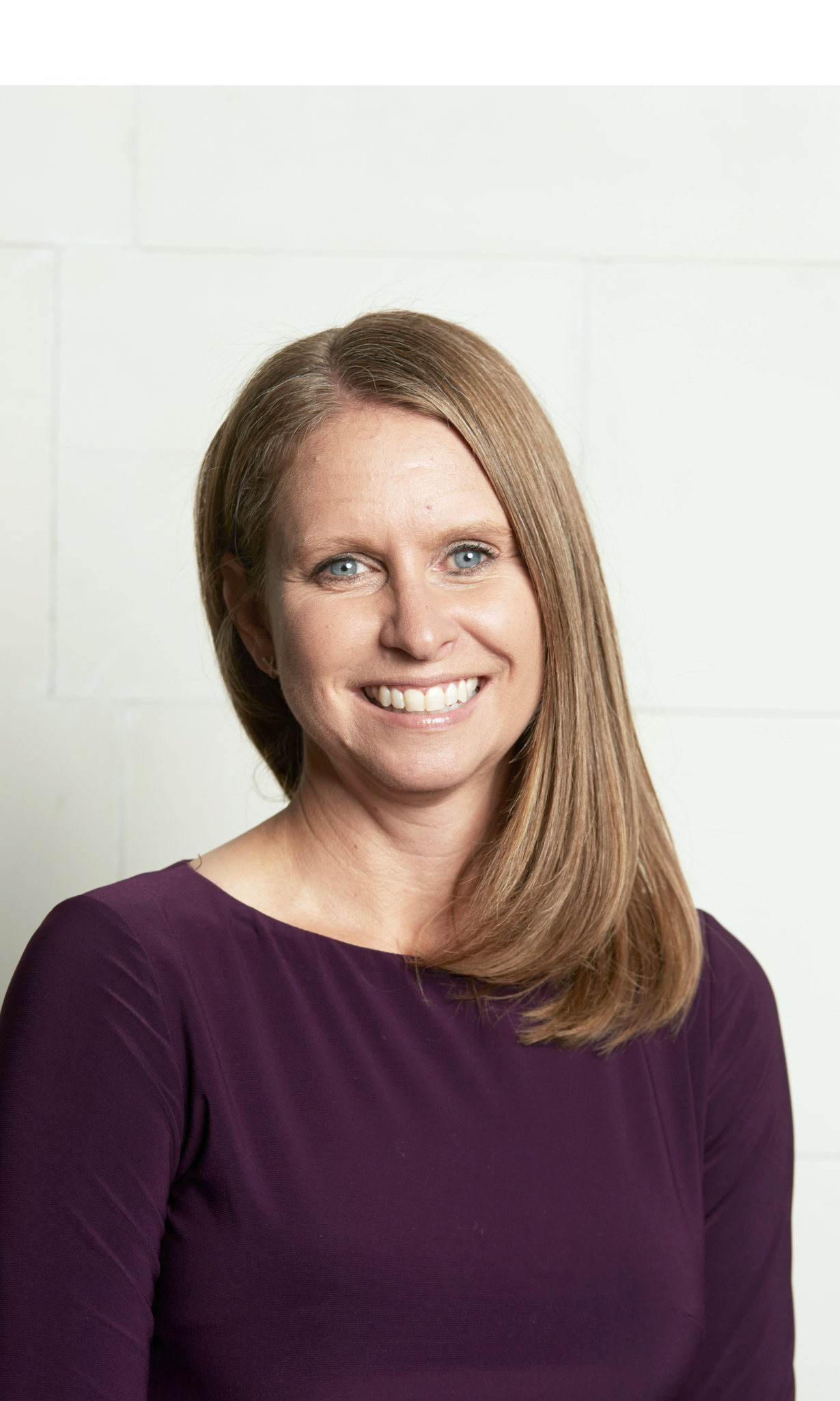 Smiling woman with shoulder-length strawberry blonde hair wearing a purple top, standing against a plain off-white wall.