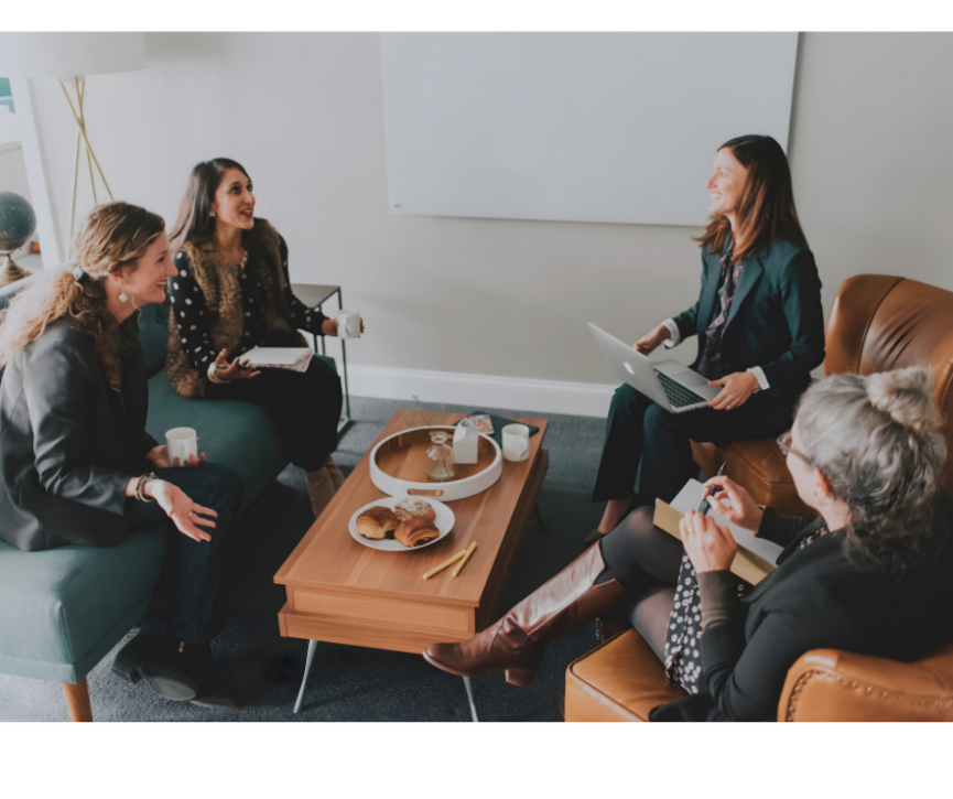 Five women sitting in a circle in a casual office, engaged in a lively discussion, with coffee mugs and pastries on the table.