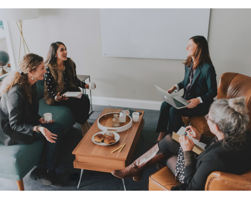 Five Inbloom Team members sitting in a circle in a casual office, engaged in a lively discussion, with coffee mugs and pastries on the table.