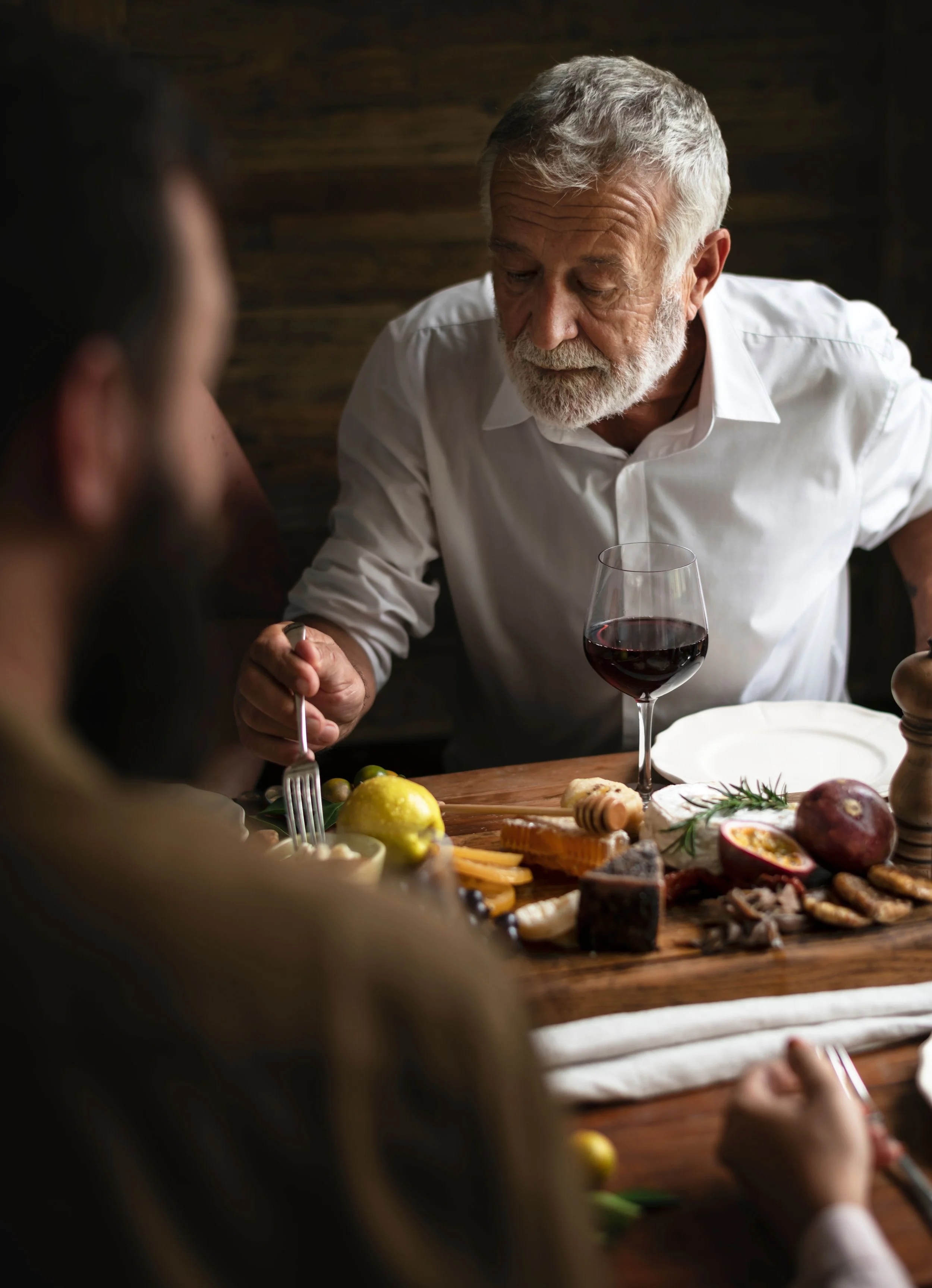 An older man with a white beard and gray hair, wearing a white shirt, eating at a table with a glass of red wine, cheese, figs, honey, and grapes.