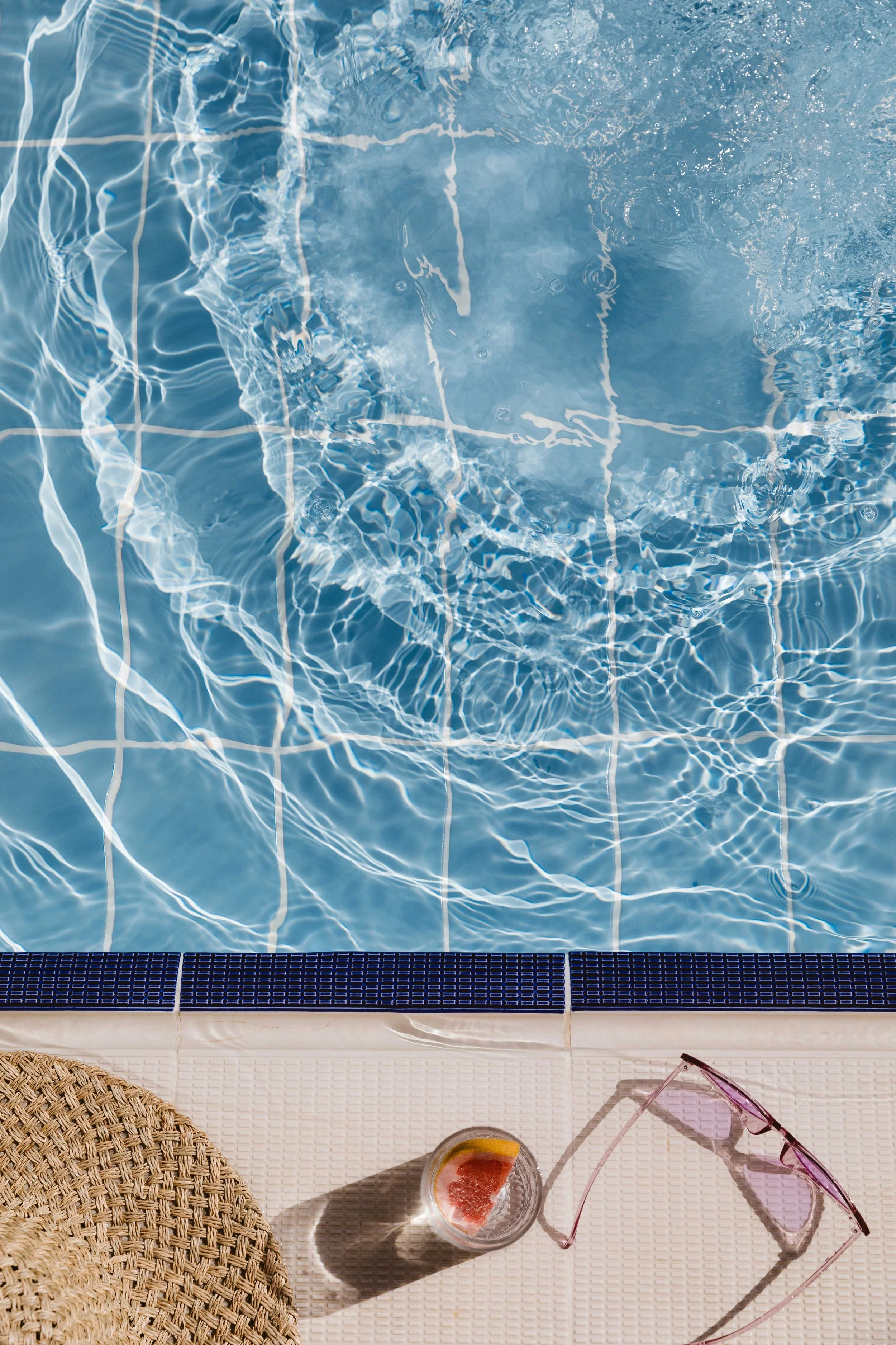 A swimming pool with clear blue water and a tiled edge, next to a white surface with a straw hat, a glass of grapefruit juice, and pink sunglasses.