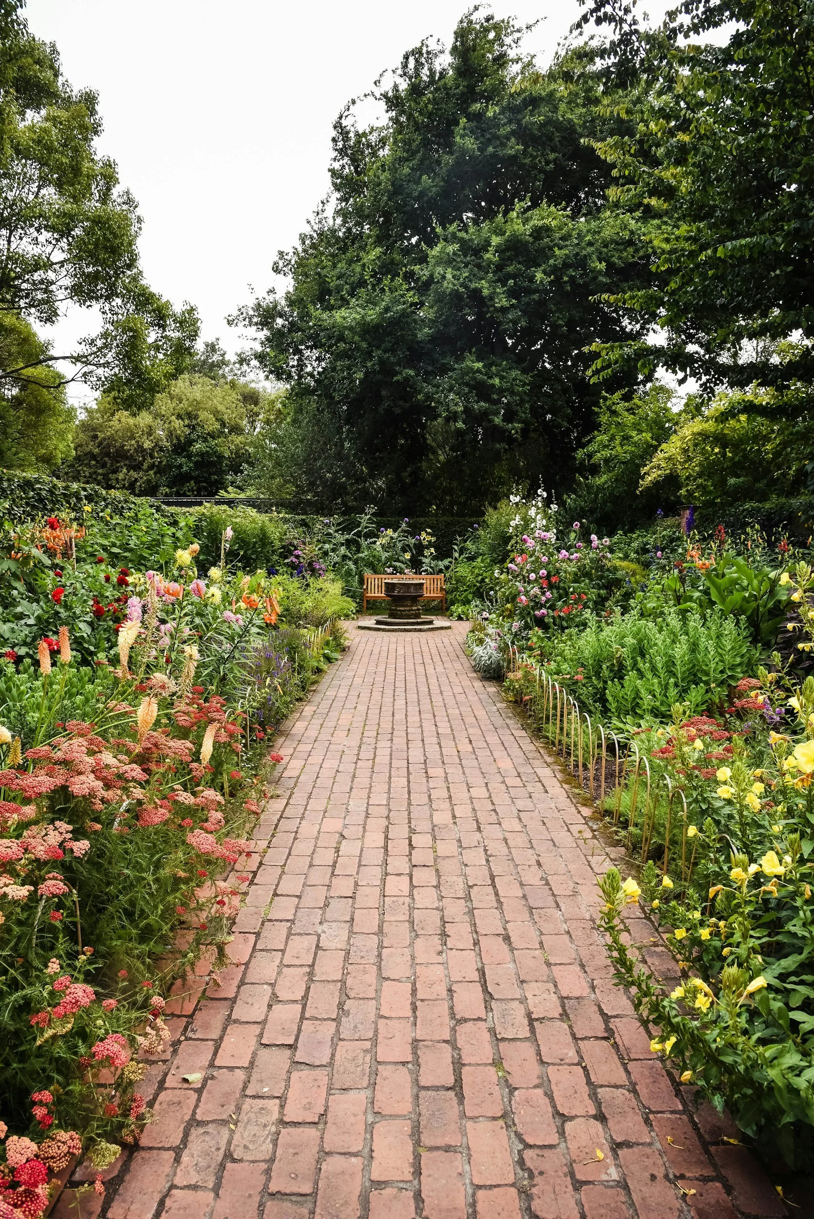 A brick pathway through a vibrant garden filled with colorful flowers on either side, leading to a wooden bench and a table in the distance, surrounded by lush green trees.