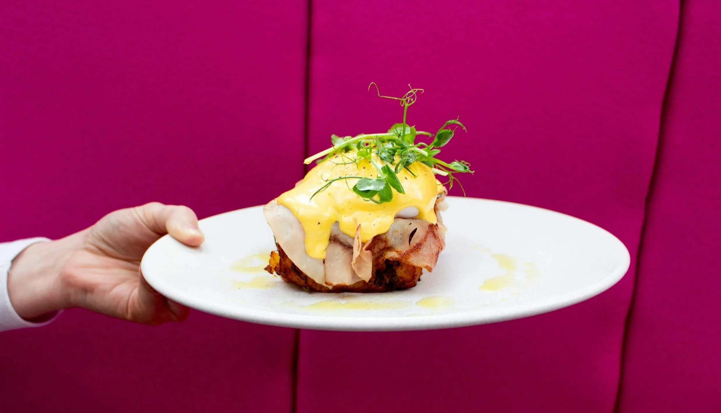 Plate with eggs Benedict topped with microgreens, served against a pink upholstered background.