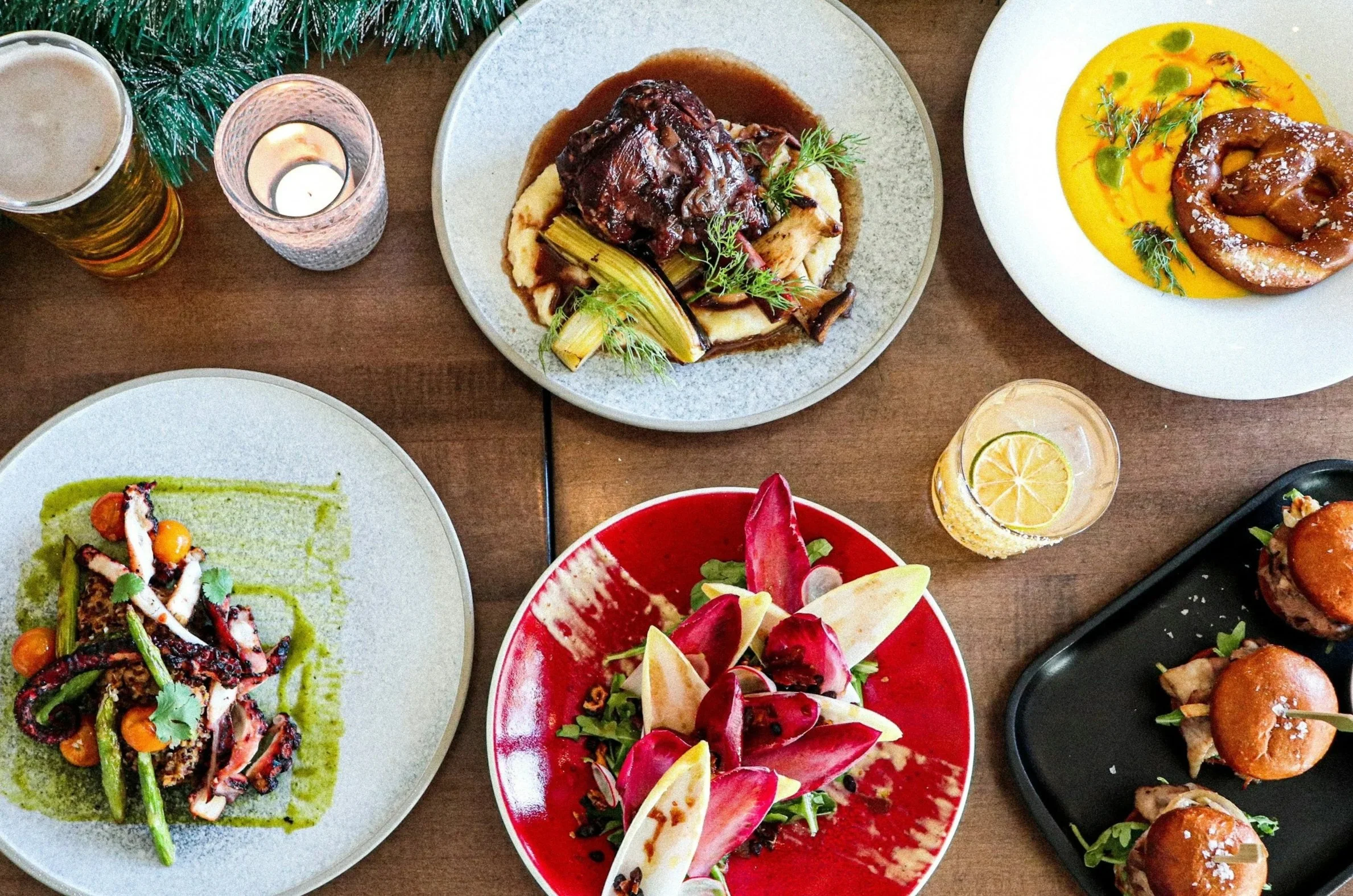 Overhead view of a table with various gourmet dishes including a roasted meat with vegetables, fish with sauce, salad with edible flowers, and sliders, along with a beer, a lemon soda, and a candle.