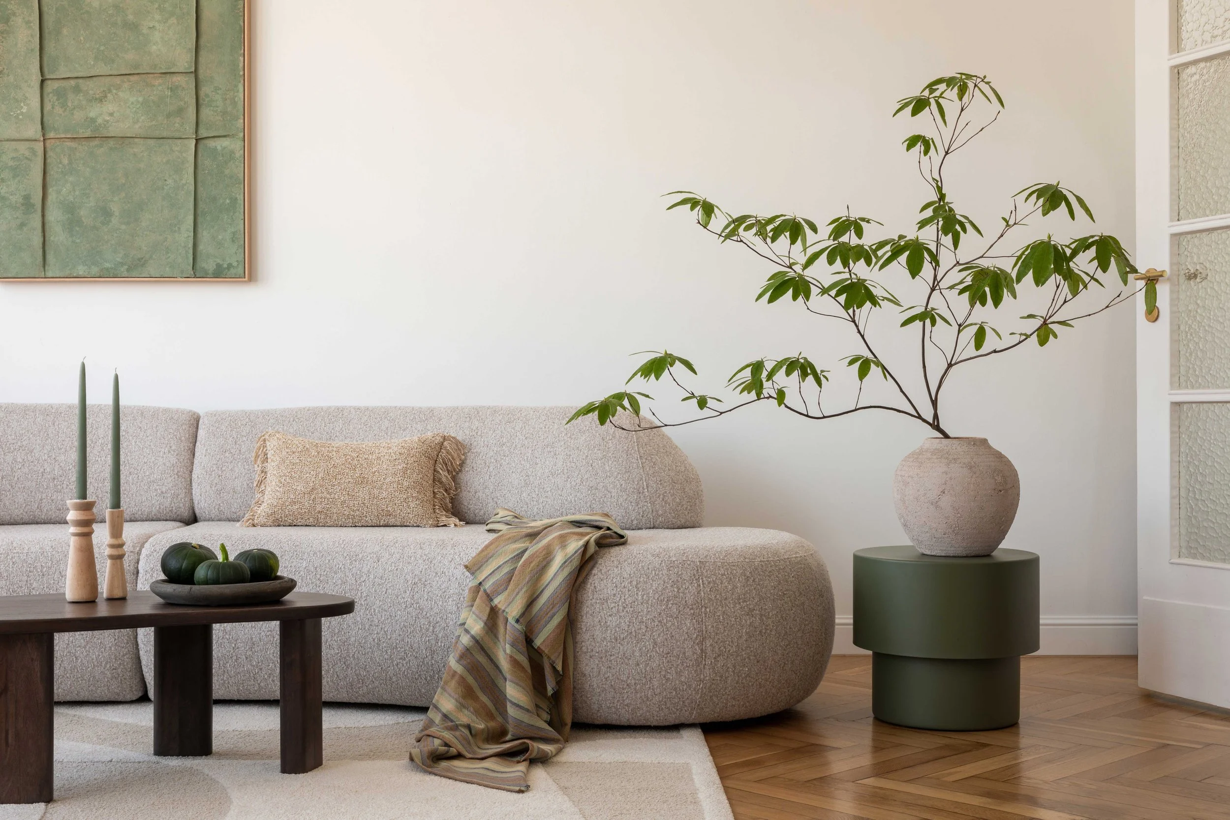 Living room with beige sofa, neutral cushion, striped throw blanket, wooden coffee table with green pumpkins and two tall green candles, large potted plant with green leaves on a green cylindrical side table, wood floor, and a framed abstract artwork on the wall.