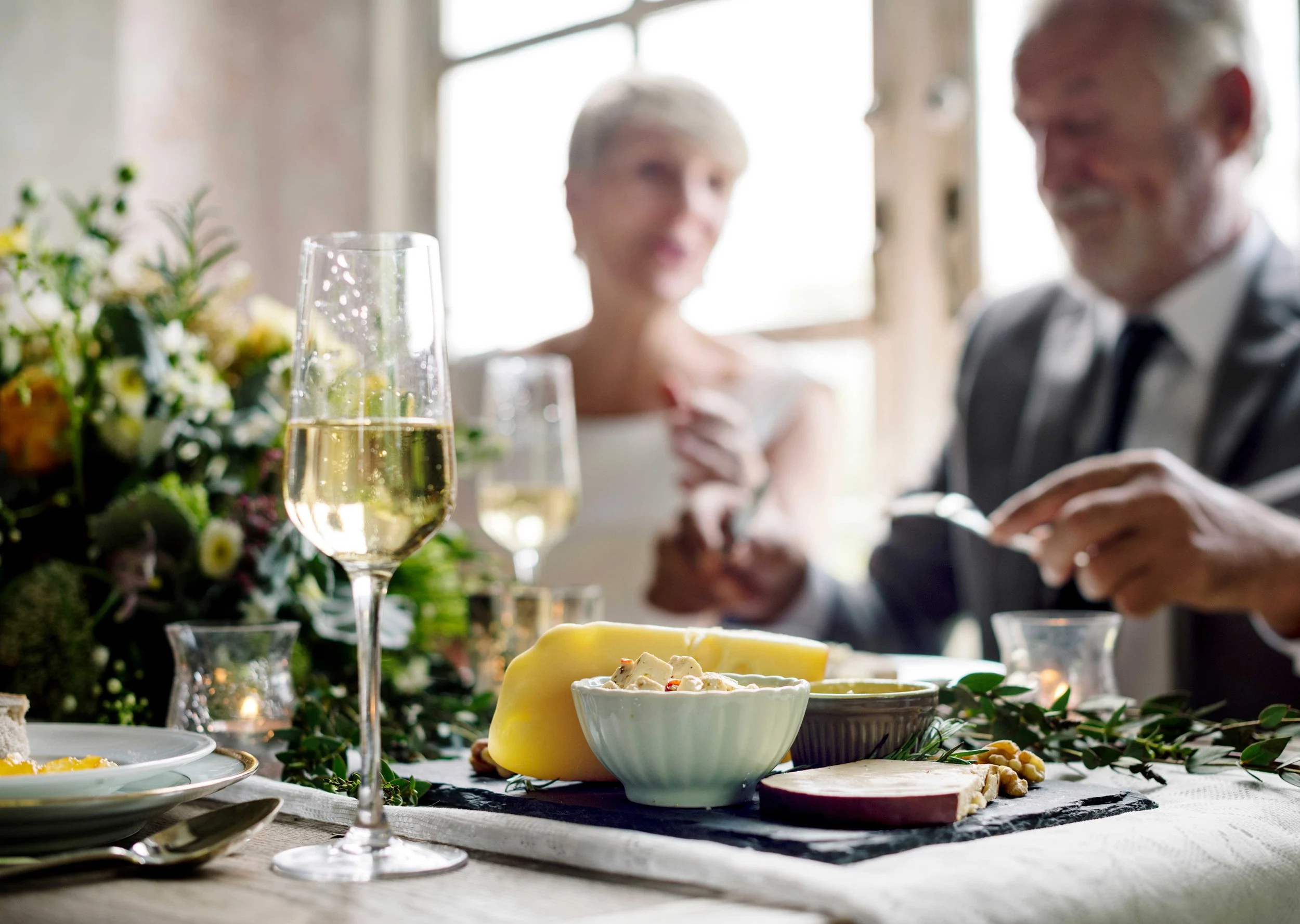 A table set for a celebration with cheese, wine, and floral decorations, with a couple in the background celebrating.