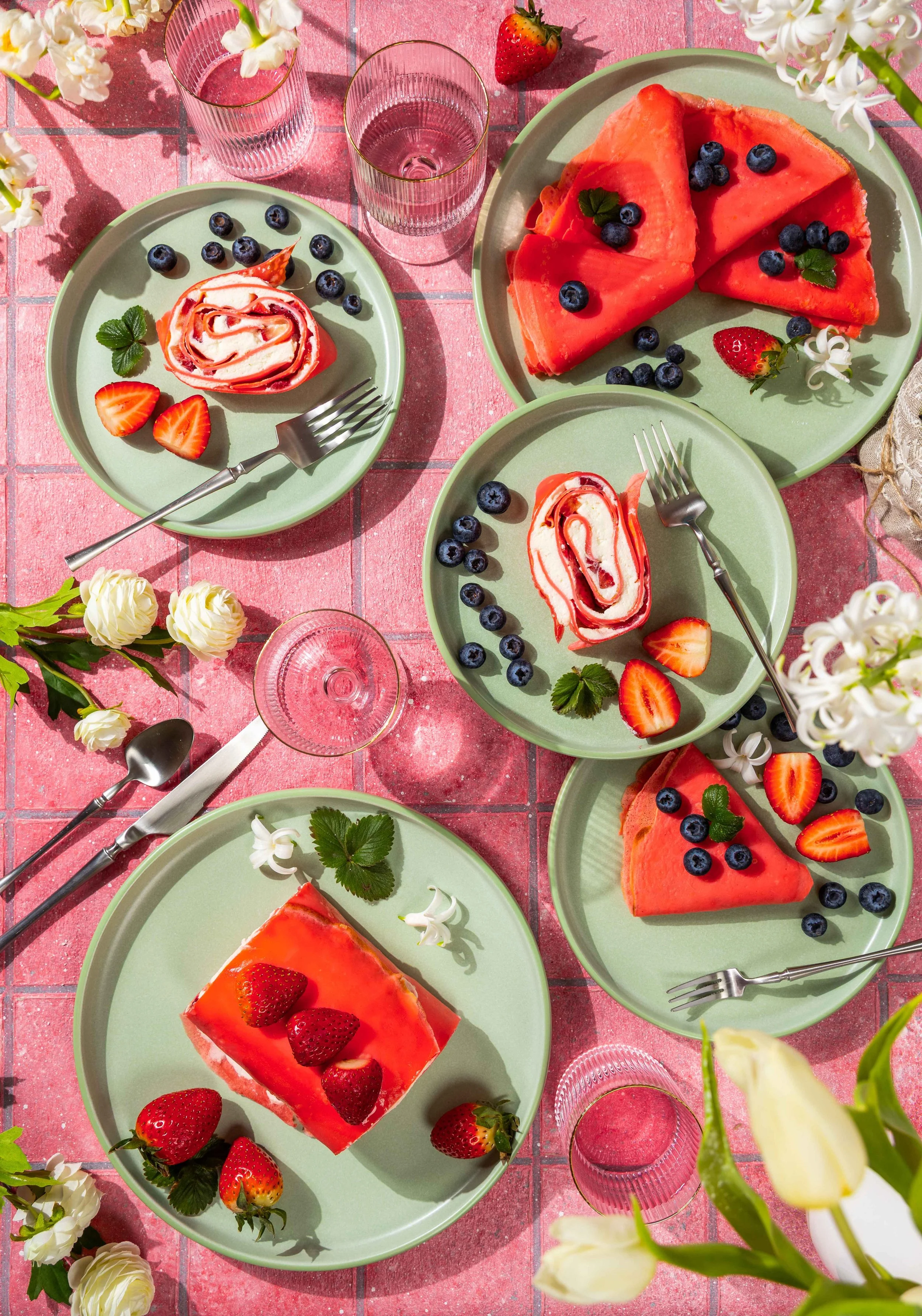 A colorful outdoor dessert table with green plates holding strawberry and berry-flavored cakes garnished with fresh strawberries, blueberries, and mint. There are several glasses of pink beverage, white flowers, and strawberries scattered around on a pink tiled surface.