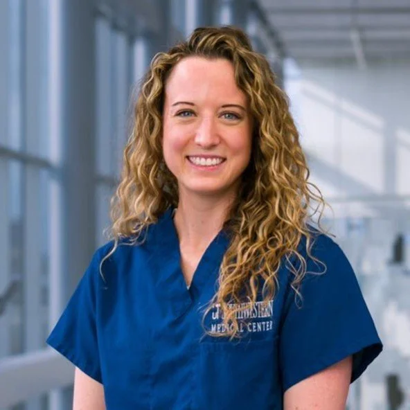 A woman with curly blonde hair wearing a blue medical scrub top, standing in a modern medical facility with large windows.