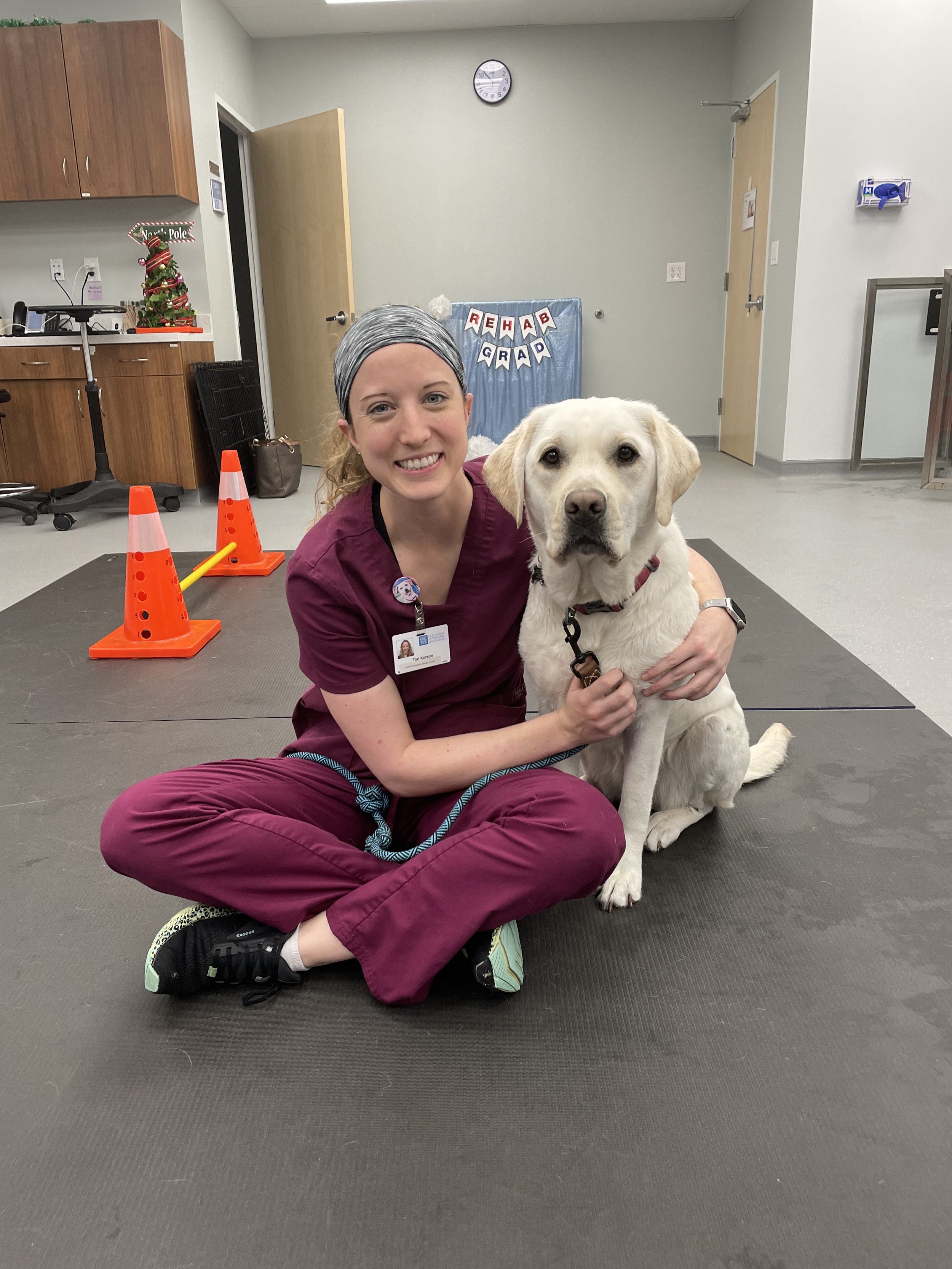 A woman in scrubs kneeling on the floor with a yellow Labrador Retriever puppy, both looking at the camera in a veterinary rehabilitation room with a sign that says 'Rehab Grad' in the background.