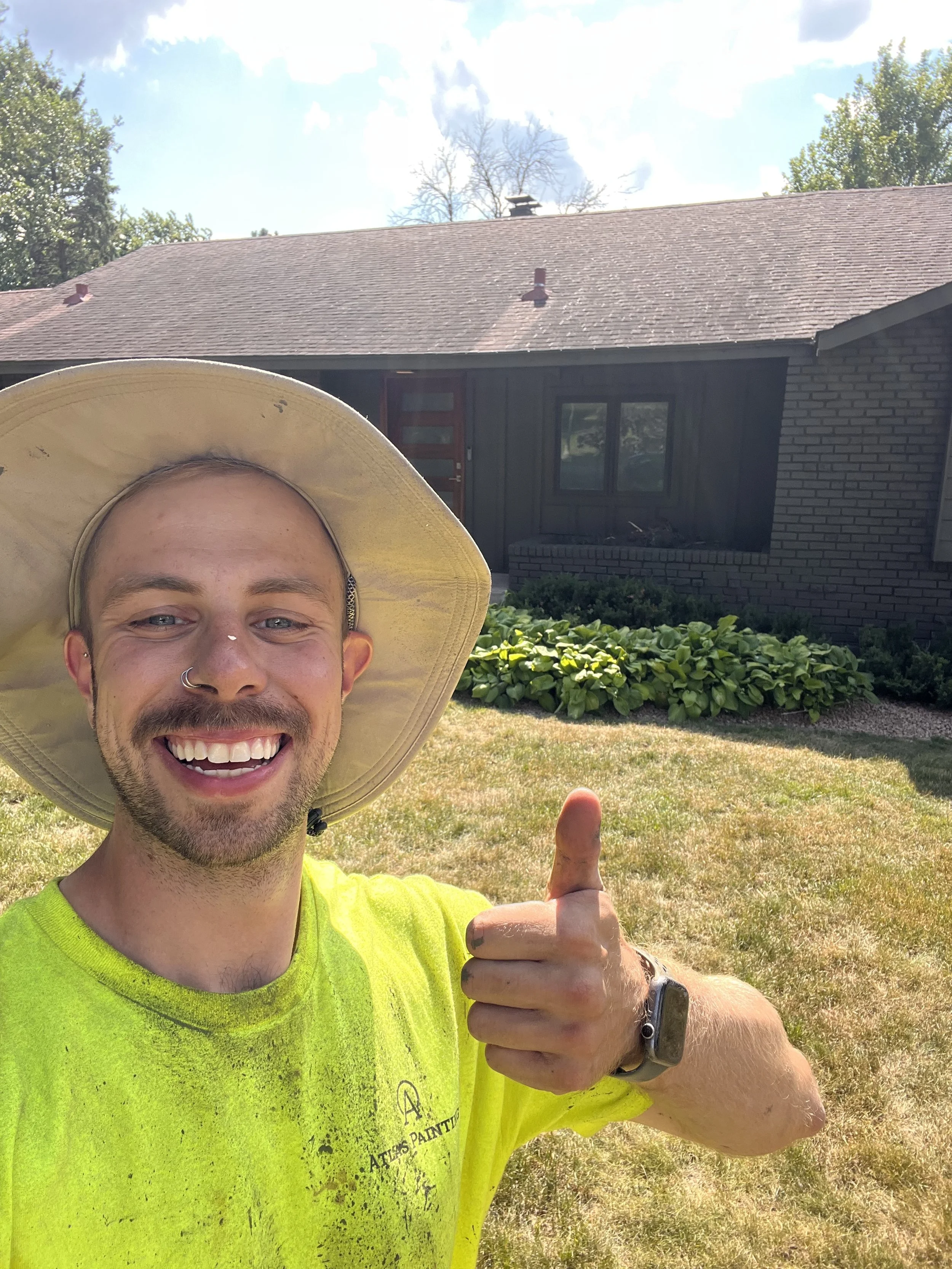 A smiling man with a beard, nose piercing, and wearing a yellow hat and bright yellow shirt giving a thumbs-up outdoors with a house, trees, and partly cloudy sky in the background.
