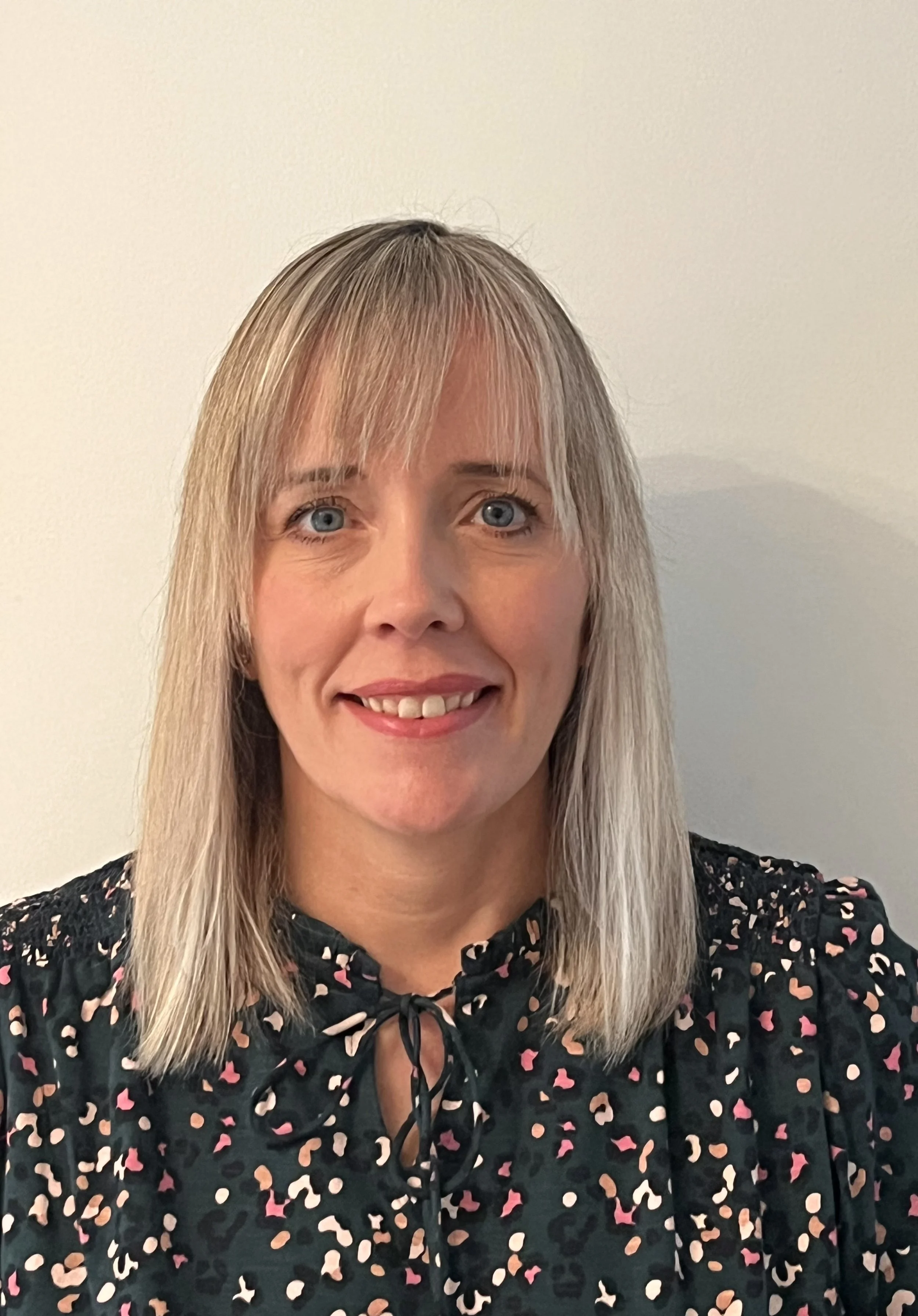Portrait of a woman with shoulder-length blonde hair, blue eyes, smiling, wearing a black floral blouse with a bow tie.